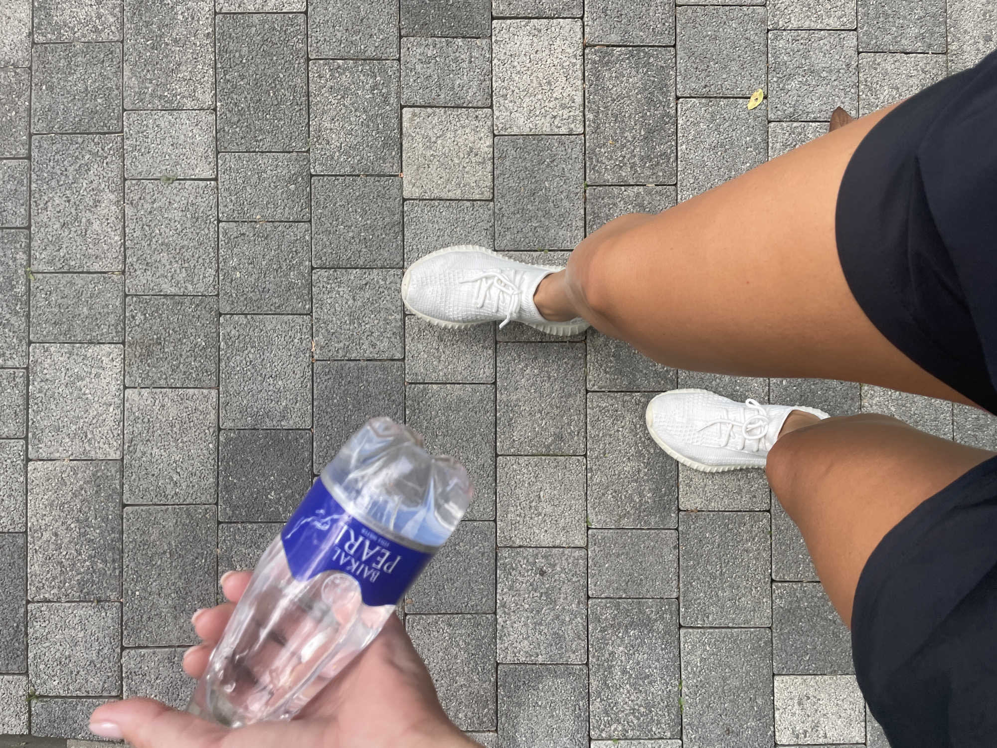 The image shows a person's legs and hand holding a bottle of Baikal Pearl water, standing on a paved walkway in Sochi, Russia. The walkway is made of gray, rectangular paving stones. The person is wearing white athletic shoes and dark shorts. The hand is holding a clear plastic water bottle with a blue and white label. The scene is outdoors, with sunlight casting shadows.