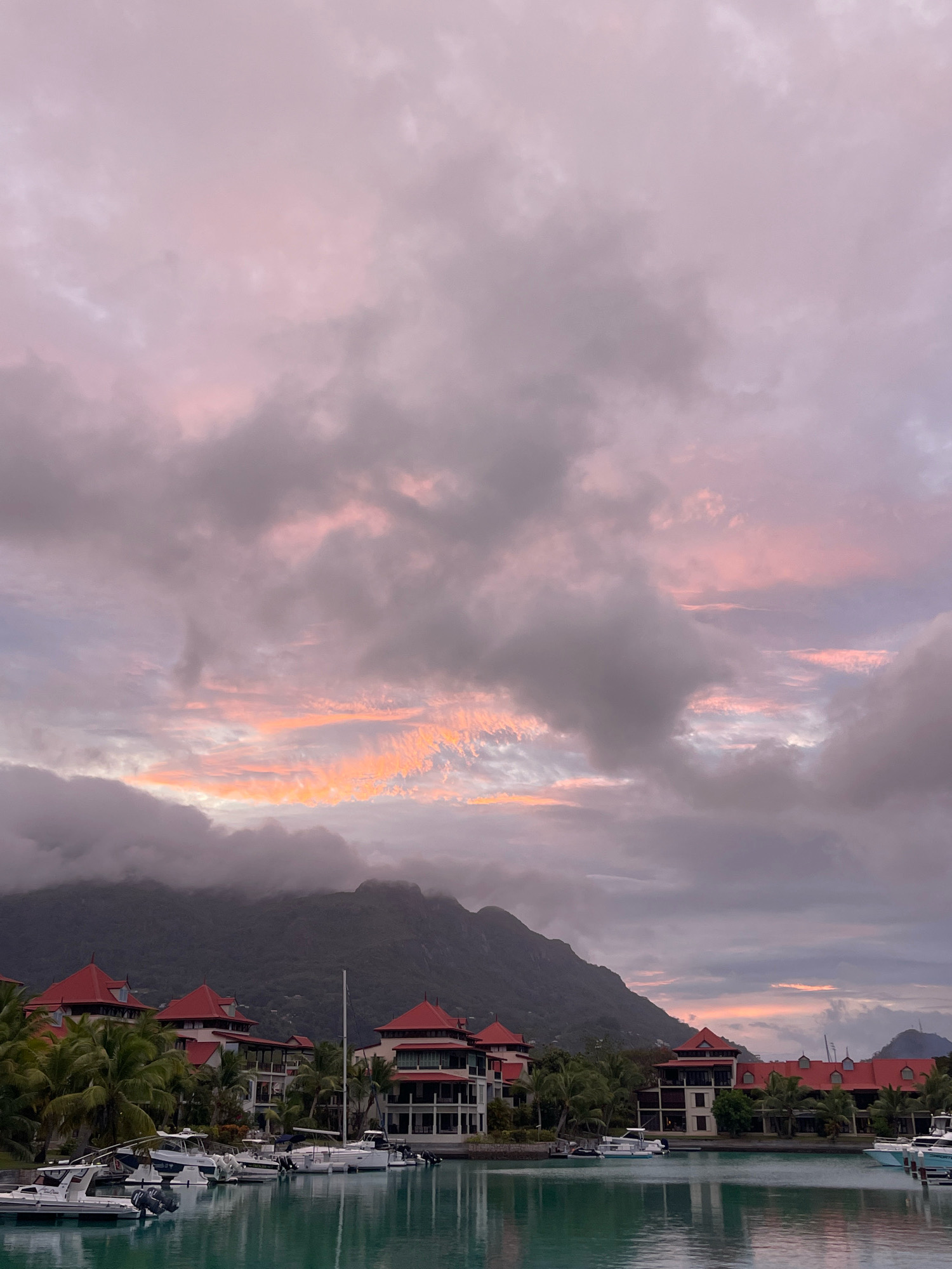 This is a photograph of a harbor with several boats docked. The buildings in the background have red roofs and are surrounded by palm trees. A mountain range is visible behind the buildings, partially obscured by clouds. The sky is filled with pink and orange clouds, indicating either sunrise or sunset. The water in the harbor is a deep turquoise color and reflects the sky and buildings. The location is Providence, Seychelles.