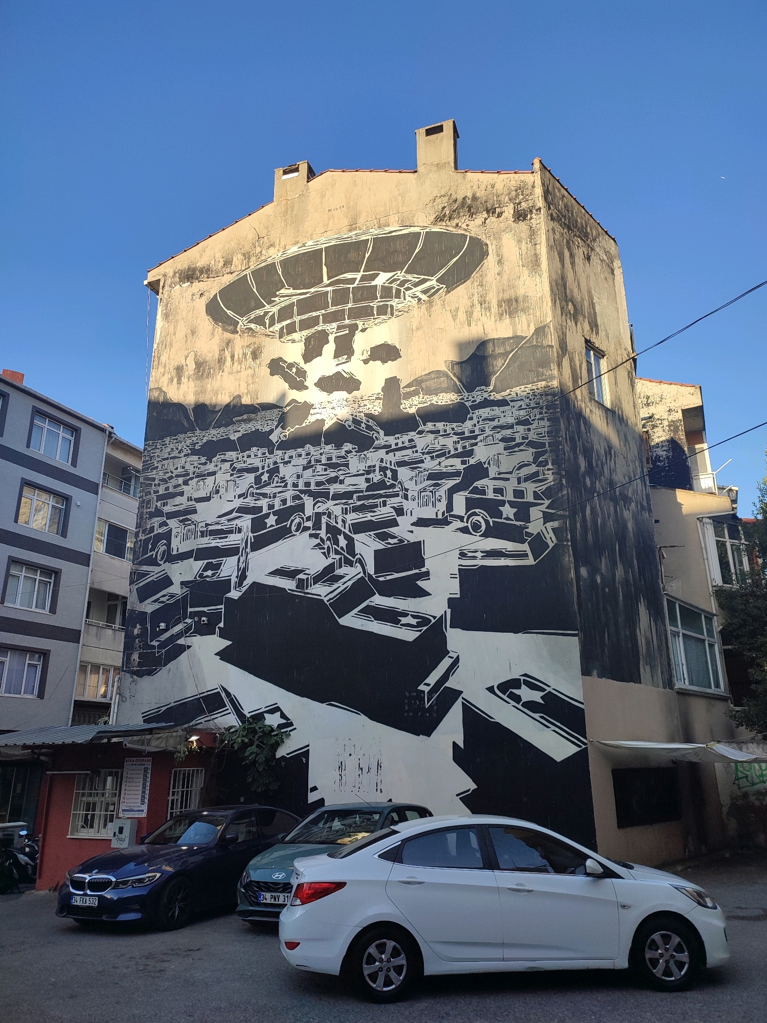 The image depicts a large mural painted on the side of a weathered, multi-story building, located in Kadıköy, Turkey. The mural, rendered in black and white, features a dystopian scene with a colossal dirigible or blimp hovering above a densely packed city landscape filled with numerous vehicles that appear to be cars and trucks. Some of these vehicles are stylized with star emblems. The overall impression is one of an airborne invasion or a crowded, chaotic urban environment.
Adjacent to the mural-covered building is a narrower, gray residential building with several windows and balconies. In the foreground, three cars are parked on a street: a dark blue sedan on the left, a light green hatchback in the middle, and a white sedan on the right. A motorcycle is partially visible to the far left. The street appears to be paved.
The buildings are situated in a built-up urban area with a clear blue sky overhead, suggesting it is daytime. Power lines are visible crisscrossing the upper portion of the frame. The mural's style is graphic and stark, emphasizing silhouettes and strong contrasts. The building with the mural shows signs of age, with patches of discolored plaster and some vertical streaks, indicating exposure to the elements. A small, red-fronted building with windows and awnings is situated at the base of the mural-covered building.