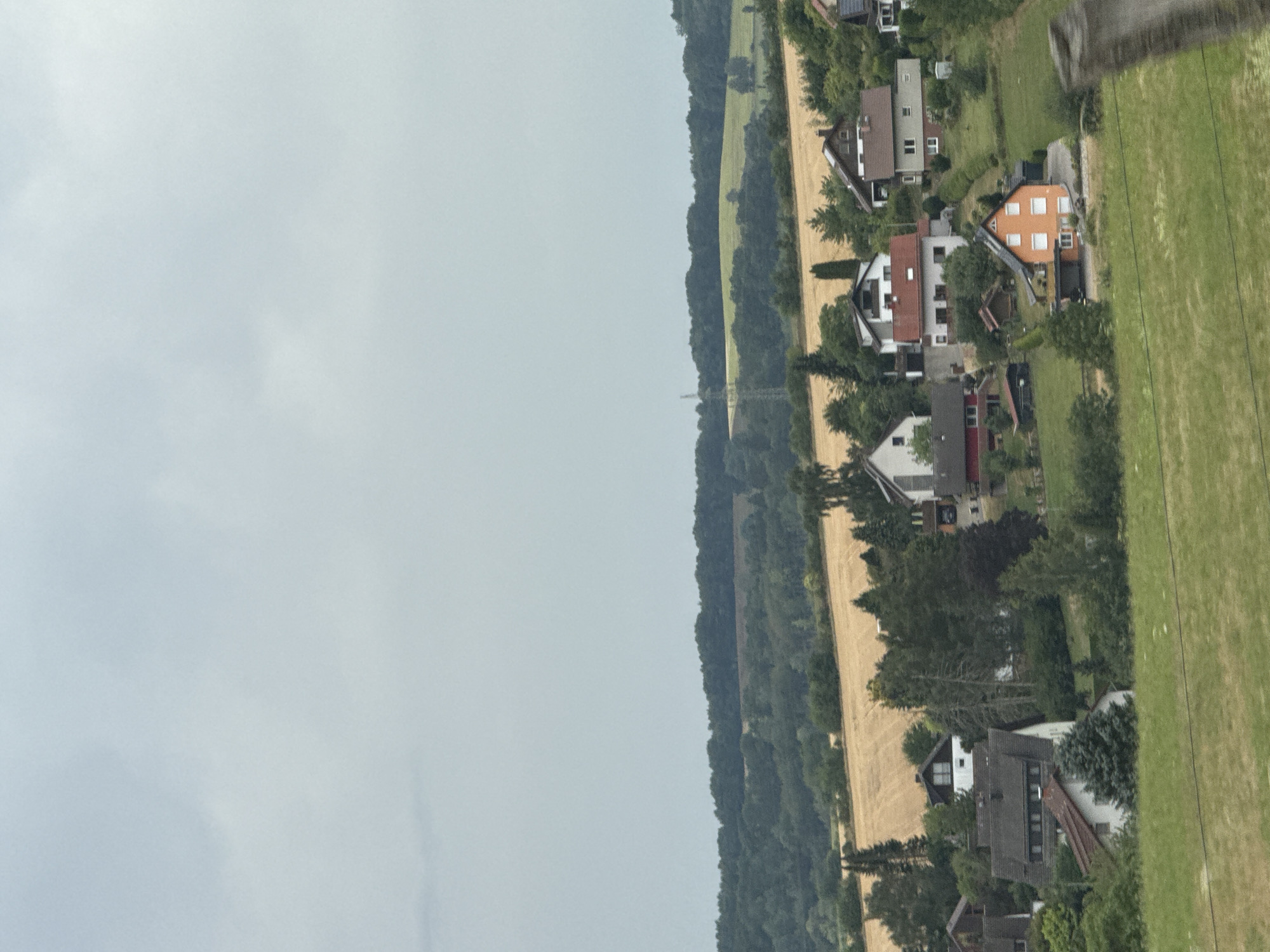 The image depicts a rural scene in the vicinity of Bad Sachsa, Germany. In the foreground, lush green grass covers a sloping hill, with a partial view of a wooden post on the right. A cluster of residential houses with pitched roofs and varying architectural styles is scattered across the midground, nestled among mature trees. The houses exhibit muted color palettes, with some featuring red or orange accents. Behind the houses, a field of dry, golden crop extends horizontally, bordered by a dense forest line. The background is dominated by a grey, overcast sky. A single electricity pylon is visible on a distant hill. There are no people or visible animals in the frame.