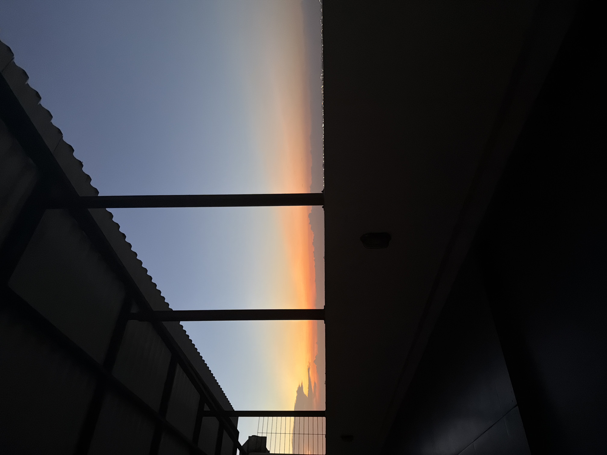 The image captures a view of a sky transitioning from blue to orange, likely during sunrise or sunset, framed by architectural structures. The perspective is from within a building or partially covered space, with a metal-framed roof structure visible on the left and the dark silhouette of an interior wall on the right. The sky is segmented into vertical bands by the roof's supports. The location is labeled as Unknown, Unknown. There are no people visible.