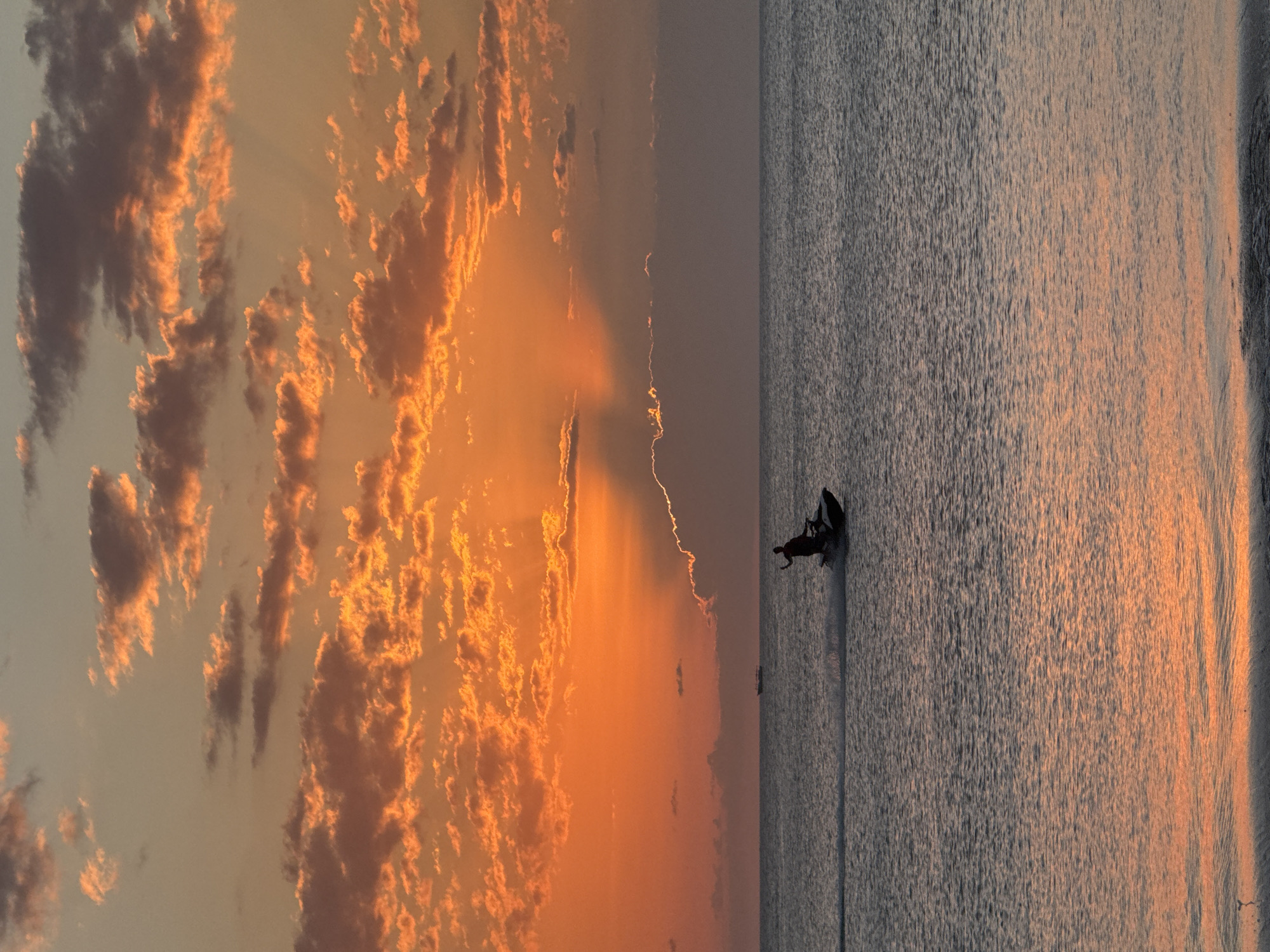An outdoor, vertical, eye-level shot shows a sunset over the ocean in Kuta Selatan, Indonesia. The sky is a brilliant orange with fluffy clouds, some of which are lit by the setting sun. The ocean is calm and reflects the colors of the sky. A person on a jet ski is in the middle of the frame, speeding across the water. There is a ship on the horizon in the background. The overall mood of the image is peaceful and serene.
