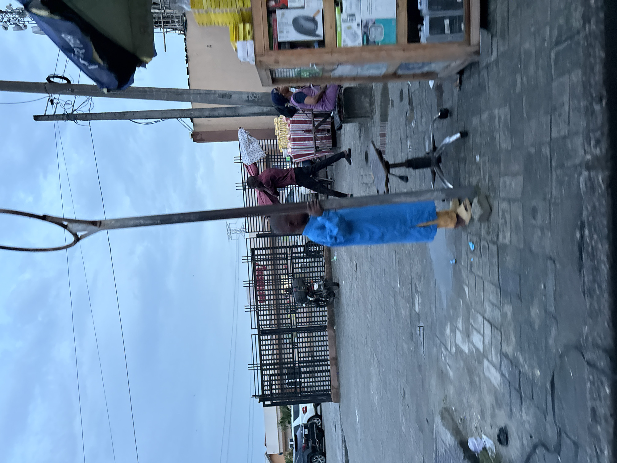 The image depicts a street scene in Ado, Nigeria, likely during overcast conditions. In the foreground, a young child wearing a blue robe and yellow sandals stands near a metal pole. Behind the child, a man in a maroon shirt and black pants carries a large red and white patterned fabric over his shoulder. To his right, a woman wearing a purple hijab and dress sits on a low bench. Adjacent to the woman is a stall with various items for sale, including what appear to be packaged goods. In the background, a black motorcycle is parked near a black metal gate. Several cars, including a white SUV and a black SUV, are visible parked outside the gate. Buildings with light-colored walls and a dark roofline are present in the distance. The ground is paved with gray interlocking bricks, and there are some scattered debris and puddles on the surface. Power lines and utility poles are visible against the cloudy sky.