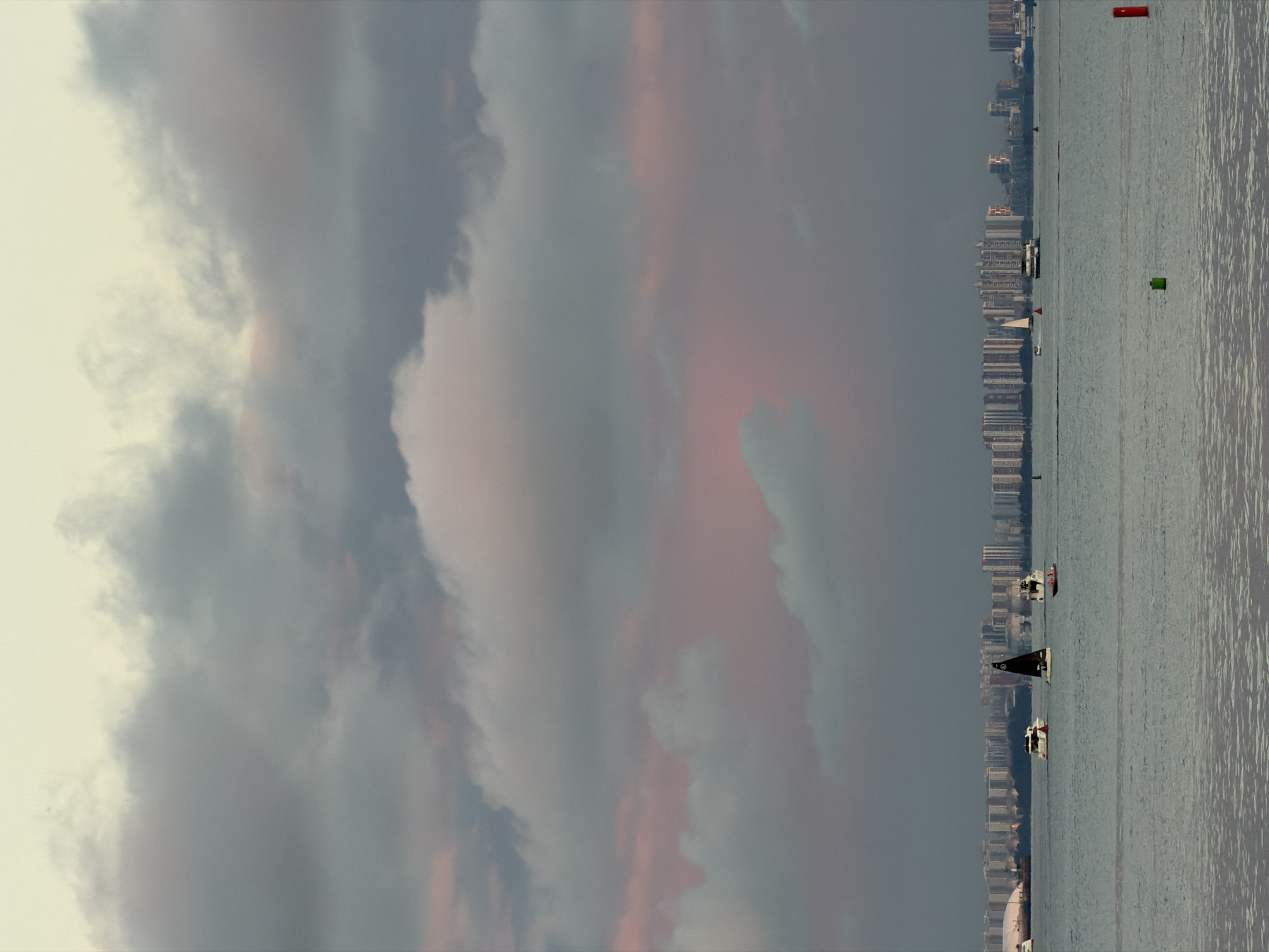 The image depicts a wide shot of a body of water with a city skyline in the background under a cloudy sky. Several boats are visible on the water. In the foreground, the water has ripples. In the middle ground, a sailboat with a black sail is prominent, accompanied by two other smaller boats. Further in the distance, a long line of buildings forms the city skyline. The sky is filled with large, dramatic clouds that are tinged with pink and gray, suggesting either sunrise or sunset. The overall scene is tranquil and hints at a coastal urban environment. The location is identified as Unknown, Russia.