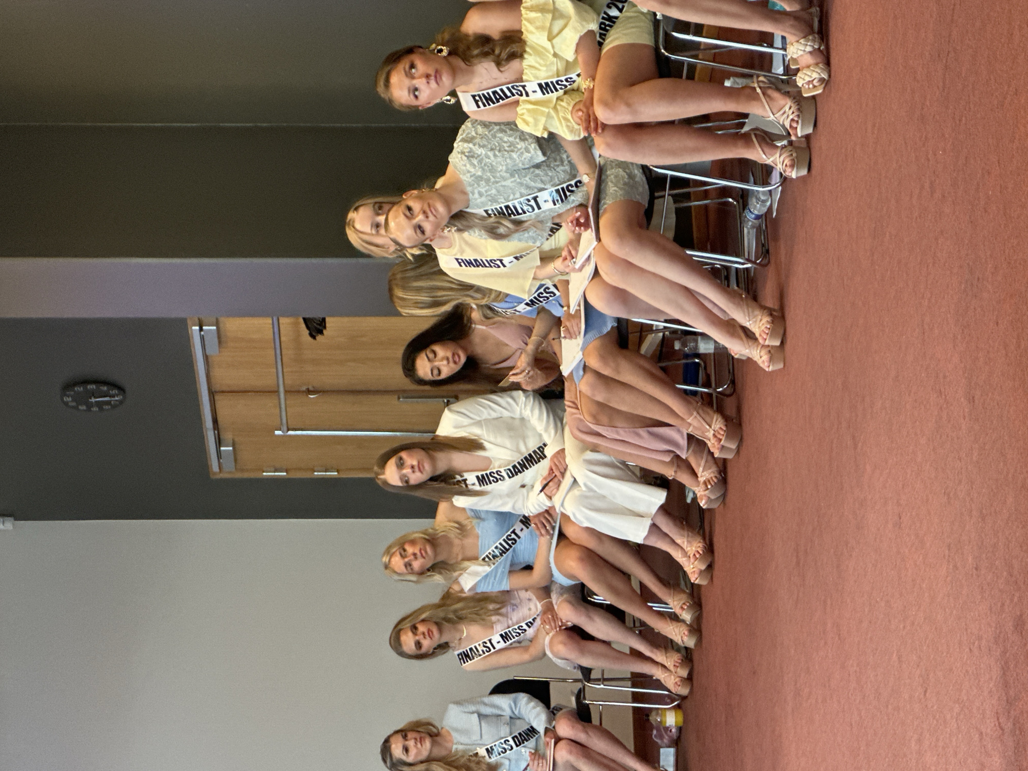 Certainly, let's analyze the image.
**Scene:** A photograph depicts a group of approximately eight young women seated in a row of chairs indoors. They appear to be contestants or finalists in a "Miss Danmark" beauty pageant, as evidenced by the sashes bearing phrases like "Finalist - Miss" and "Miss Danmark." The location is unknown.
**People:**
* Eight young women of various hair colors (blonde, brunette) and skin tones are present. They are dressed in a variety of light-colored dresses and skirts, mostly in pastel shades (yellow, blue, white). They are wearing heeled sandals. They all wear the same sash.
**Objects:**
* Chairs
* Sashes
* A water bottle
* Carpet
**Actions:**
* The women are seated.
**Environment:**
* Indoor setting.
* The wall is white with gray panels.
* A wooden door is partially visible on the left.
* There is a clock on the wall.
* The floor is covered with red carpet.