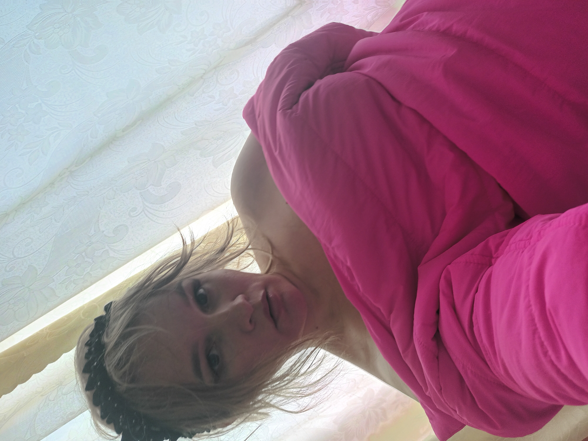 The image presents a blonde-haired female, appearing to be in her 30s-40s, situated in an indoor environment in the city of Ольховка, Russia. She is depicted from a low-angle perspective, wrapped in a large, fuchsia-colored blanket or comforter that covers her shoulders and torso, leaving parts of her upper chest exposed. A black headband, possibly with floral or feather-like embellishments, is worn on her head. Her expression is neutral, with a direct gaze toward the camera. The background consists of light-colored curtains, likely white or cream, adorned with a subtle embossed floral pattern, suggesting an interior space with a window.