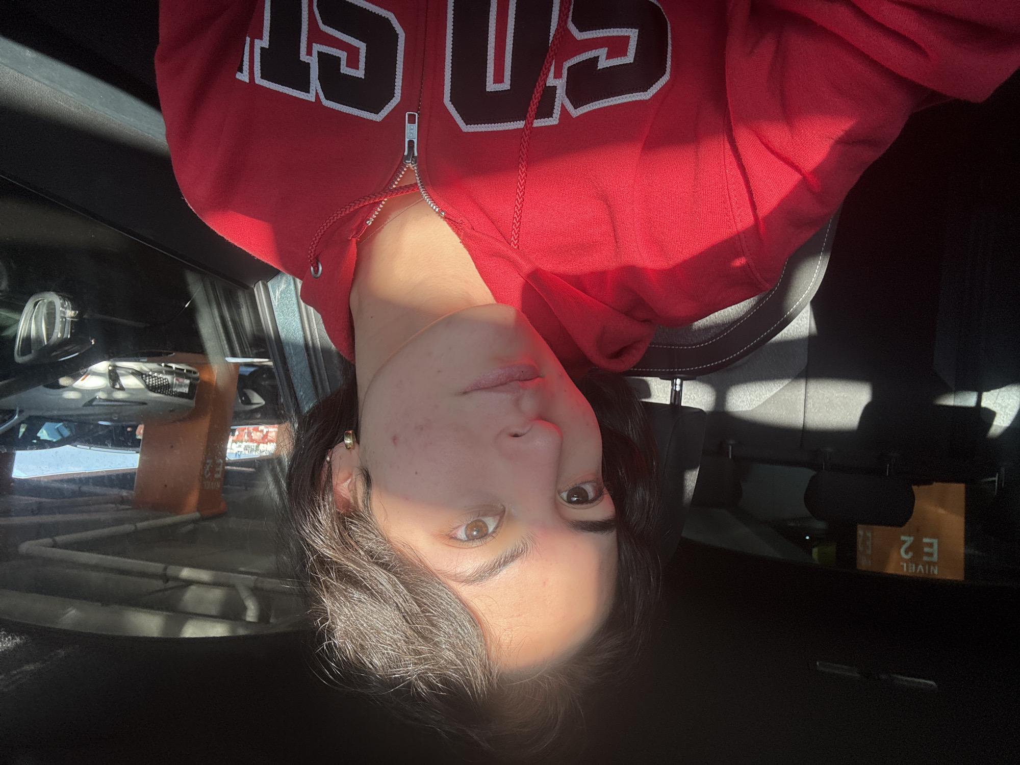 An image, originally oriented upside down, depicts a young person from the chest up, positioned inside a vehicle, likely a car. The person is wearing a red zip-up hoodie with white and black letters on the chest, which appear to spell "GS" or "CS." Strong, natural sunlight illuminates the right side of their face and torso, creating a prominent shadow on the left side of their face and body. The person has dark hair and a neutral expression, looking directly at the camera. Their right eye, illuminated by the sun, appears a lighter brown/amber, while their left eye is in shadow and appears darker. A small gold hoop earring is visible in their right ear.
Through the car window on the left, an orange traffic cone is visible, along with parts of what appears to be a structure or building in the background. In the upper right corner, a rear-view mirror reflects an inverted orange sign displaying the text "E 2" and "NIVEL." The scene suggests it is daytime, possibly in a parking area. The location context provided is San Bernardino Tlaxcalancingo, Mexico.