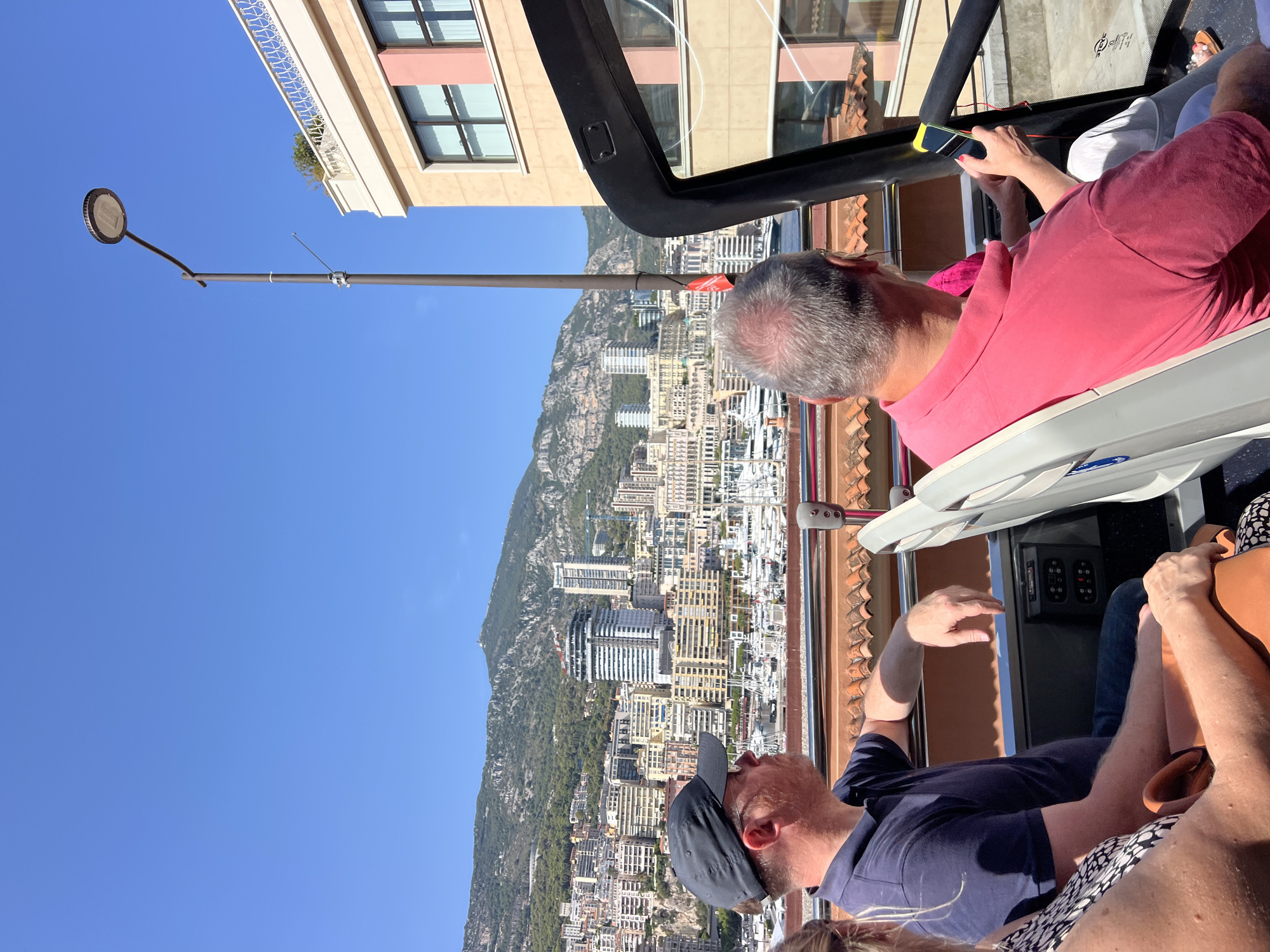 In Monaco, on a sunny day, people are seated on the upper deck of an open-top bus, a tour bus, overlooking a harbor filled with yachts and a dense city built on a hillside. The city is characterized by numerous buildings of varying heights and architectural styles, tightly packed together, reaching up towards a majestic mountain range in the background. The bus passengers, facing away from the camera, are engaged in observing the scenery. The foreground shows the backs of two men and a woman, with one man in a navy polo shirt and baseball cap and the other in a pink polo shirt holding a smartphone. The bus's black frame is visible, as is a lamppost to the right. The environment is clearly urban and coastal, with the Mediterranean Sea implied by the presence of the harbor and yachts.