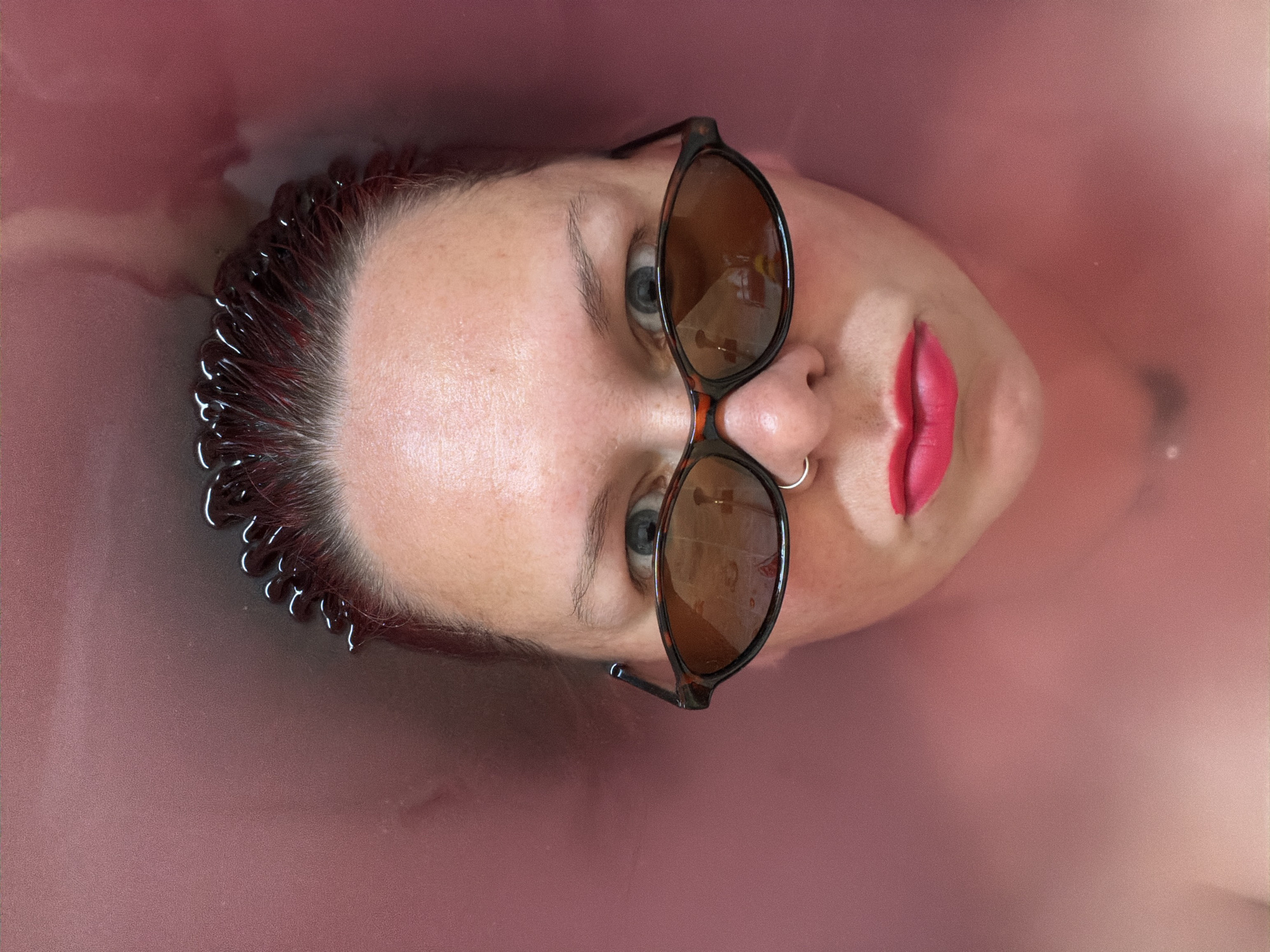 A woman's head is tilted back in a tub of pink water. She wears sunglasses with brown lenses and frames, and bright red lipstick. Her hair is dark at the roots and red-tinged at the ends, with water beading along the hairline. She has pale skin and blue eyes. A silver ring pierces her left nostril. The background is a uniform pink color of the water in the tub. Location: Unknown, Unknown.
