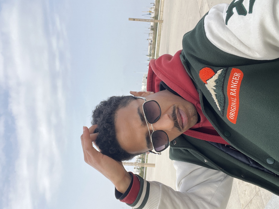 A single adult male is depicted in a half-body outdoor portrait. He has dark, curly hair, a neatly trimmed mustache, and wears aviator-style sunglasses with gradient lenses. He is dressed in a red hooded sweatshirt layered under a dark green varsity jacket with white sleeves. The jacket features an embroidered patch on the left chest showing a mountain, a red sun, and the text "ORIGINAL RANGER". His right hand is raised, with fingers lightly touching his hair.
The setting is an open, paved waterfront area, identified as a promenade or pier along the marina or port in Tanger طنجة, Morocco. In the mid-ground and background, numerous boat masts from a marina are visible, along with distant cranes and buildings lining the coastline. The sky is overcast with scattered clouds, providing diffused natural light. The ground is light-colored pavement.