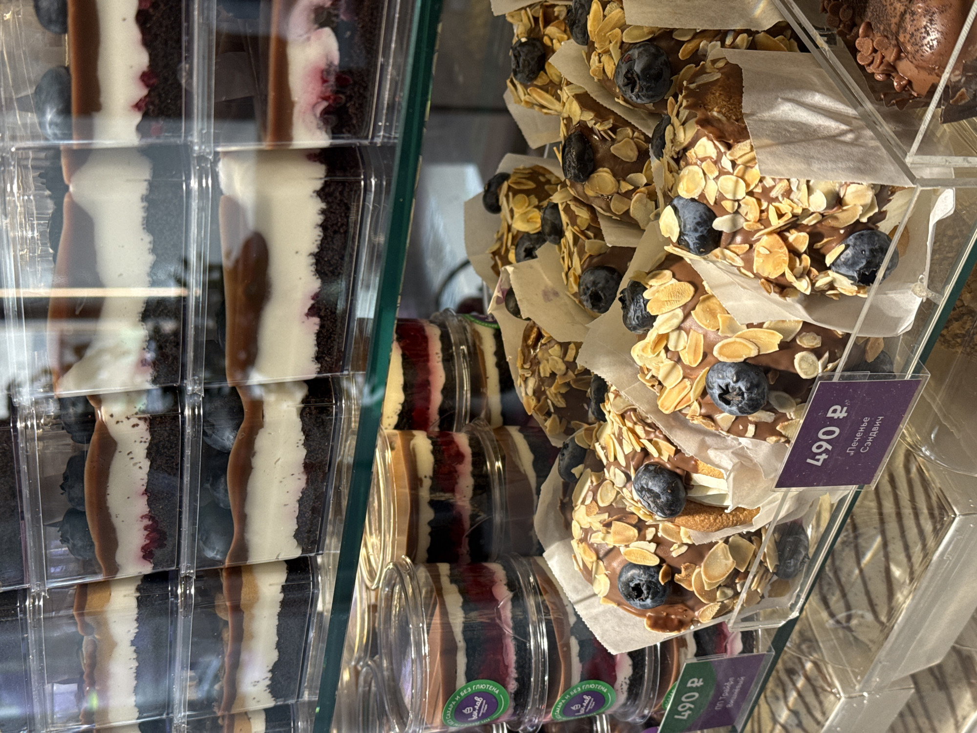 The image shows a display case filled with various desserts in what appears to be a bakery or cafe in Krasnoyarsk, Russia, as indicated by the price tags and text in Russian. On the left are layered desserts, likely trifles or mousse cakes, packaged in clear rectangular containers. The layers consist of dark cake or cookie crumbs, light-colored mousse or cream, and what appears to be fruit filling. Some of the cakes are garnished with blueberries.
To the right are what the labels describe as "Peченье Сэндвич" (cookie sandwich) or some type of layered donut snacks. These are round and coated with chocolate and sliced almonds, also topped with blueberries and wrapped in parchment paper. They are stacked on top of one another. Price tags indicating "490 P" (Russian Rubles) for both the rectangular dessert cups and the cookies. The setting suggests a modern, well-lit food establishment.