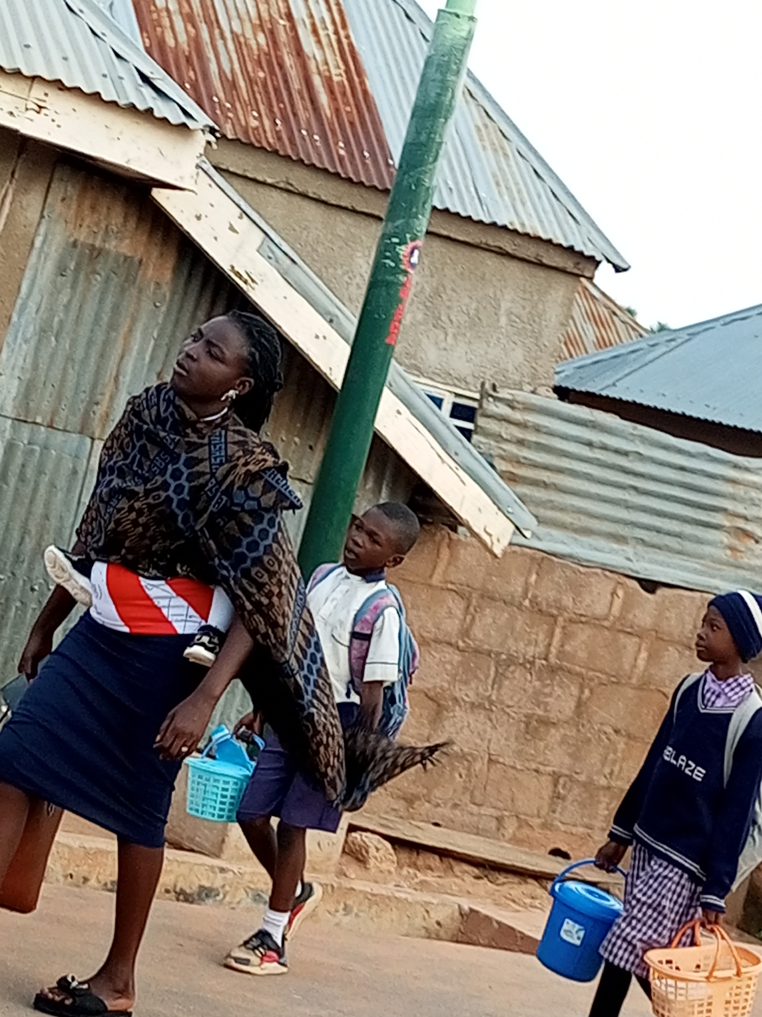In Gombe, Nigeria, a woman carries a child on her back, wearing a patterned wrap and dark skirt. Beside her, a young boy in a white shirt and backpack walks, while another child in a school uniform with a blue sweater that reads "BLAZE" and a plaid skirt carries a blue bucket and a basket. They are walking on a dirt path next to a brick wall. In the background, corrugated metal roofs and buildings are visible, suggesting a residential or commercial area within the city.