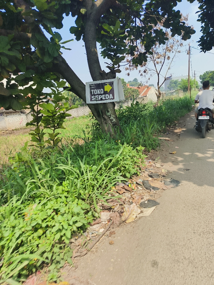 The image shows a dirt road in Cileunyi, Indonesia, at the coordinates -6.947 latitude and 107.726 longitude. A sign on a tree indicates a "TOKO SEPEDA" (bicycle shop) to the right. A man in a blue shirt rides a motorcycle away from the camera down the road. The motorcycle is black, has a license plate with the number D 6231 AAE, and a yellow reflective triangle on the back. There are a few houses visible in the background and dense green vegetation on either side of the road. The dirt road is lined with debris and scattered leaves. The sky is a bright blue with some white clouds.