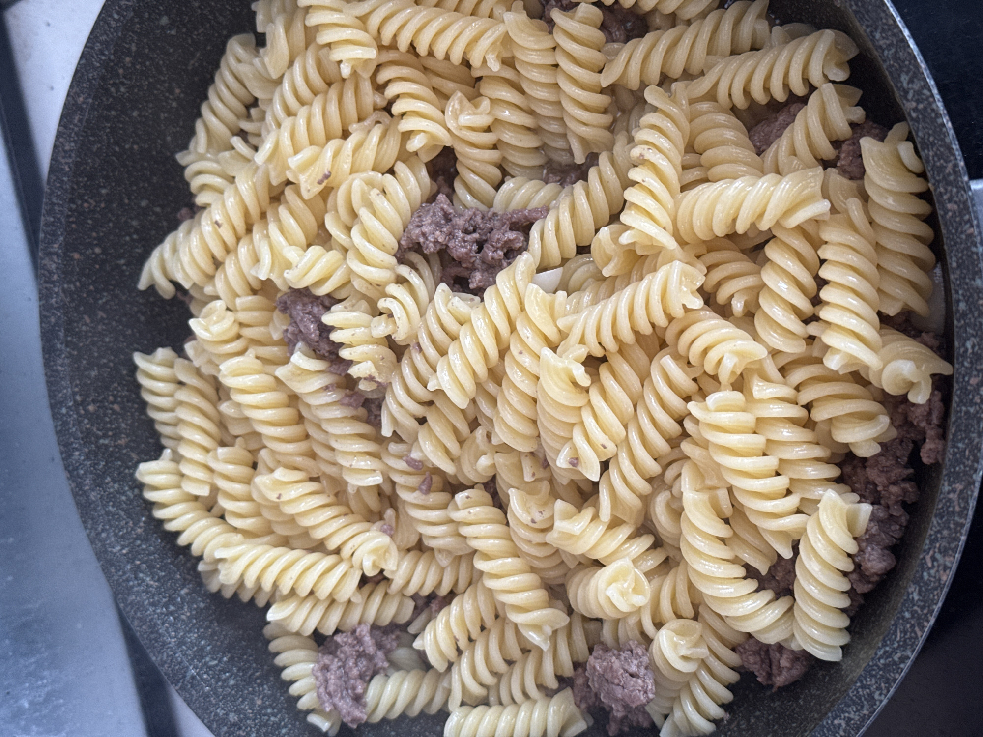 A close-up shot shows fusilli pasta mixed with ground meat in a dark gray, speckled pan. The pasta is a pale yellow and appears to be cooked al dente, with visible spiral shapes. The ground meat is a dark brown, cooked, and crumbly texture, interspersed among the pasta spirals. The pan has a textured rim and is placed on a dark surface, possibly a countertop or stovetop. The lighting is bright, casting subtle shadows within the crevices of the pasta. The image is taken from a top-down perspective, focusing solely on the food within the pan. There are no visible people or specific architectural elements that would indicate a precise location in Khimki, Russia.