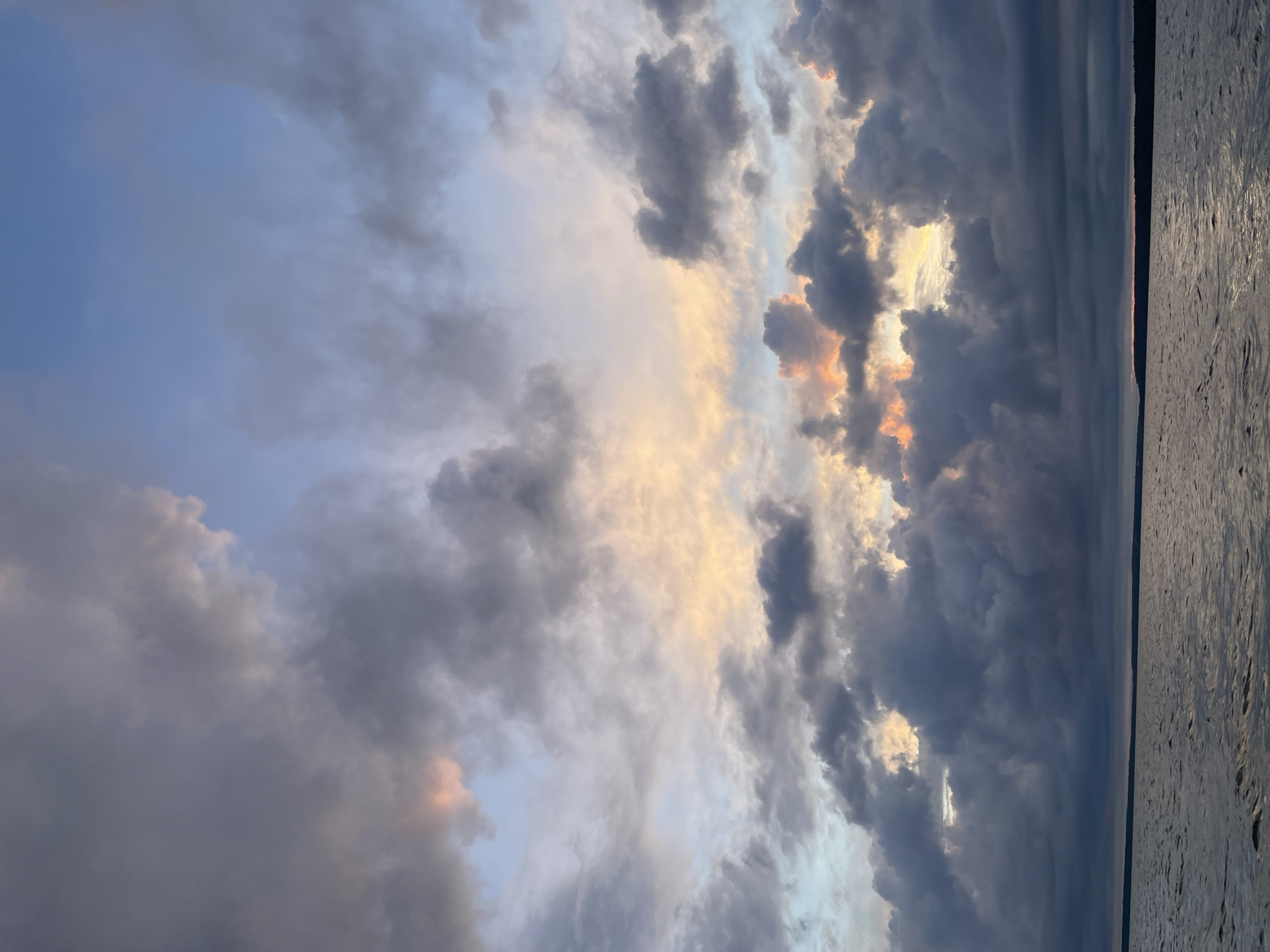 The image shows a body of water, likely a lake or river, under a dramatic sky filled with clouds. The water's surface is rippled, reflecting the muted colors of the sky. Dark, heavy clouds dominate the lower half of the sky, while lighter, more scattered clouds with hints of orange and yellow can be seen breaking through in the upper half. The horizon line is visible, separating the water from a dark silhouette of land, possibly trees or a distant shoreline. The overall impression is one of a brooding, perhaps approaching, storm or the end of a day. No people or specific structures are visible that would indicate a location within Yablonovka, Russia.