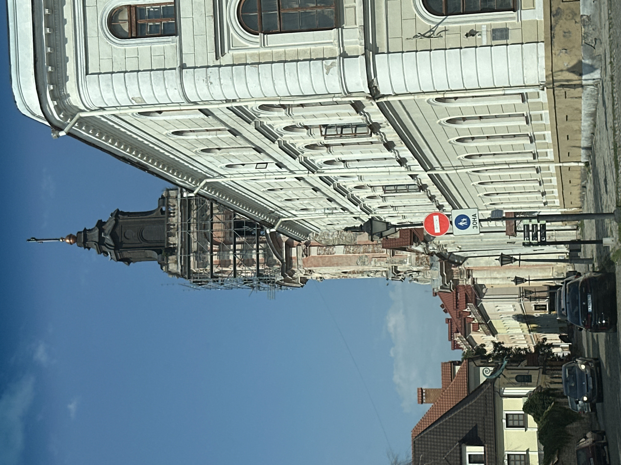 This image captures a street view in Kamianets-Podilskyi, Ukraine, dominated by historic architecture. On the right, a large, light-colored building with arched windows and ornate detailing runs along the street. To the left, a tall, dark tower, appearing to be under renovation with scaffolding, rises against a clear blue sky. The tower has a complex architectural design, including a spire at its peak. In the foreground, a street with cobblestones is visible, with several cars driving or parked. On the left side of the street, traditional buildings with red tile roofs are present. A street sign indicating a "30 ZONE" and a pedestrian crossing symbol is visible on the right. The overall scene suggests a well-preserved historical city center.