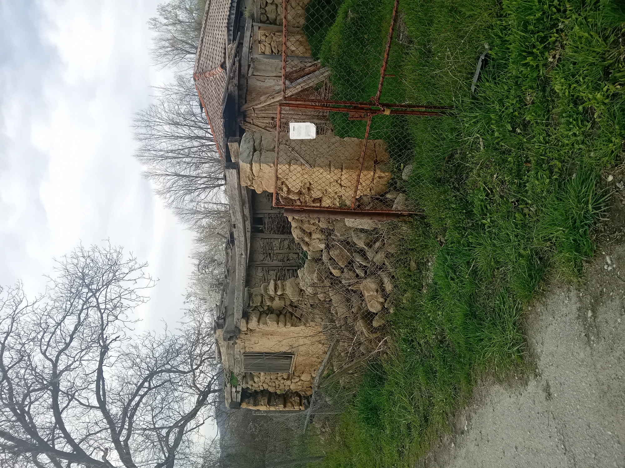An exterior shot depicts a dilapidated stone and wood building with a rusty chain-link fence in front. The location is likely rural, with grass growing along the fence line and a dirt path visible. The building is in a state of disrepair, with crumbling stonework and rotting wood. The fence is rusty and partially obscured by vegetation. Bare trees are visible in the background, under an overcast sky. A small white sign is attached to the fence. The specific location is unknown.