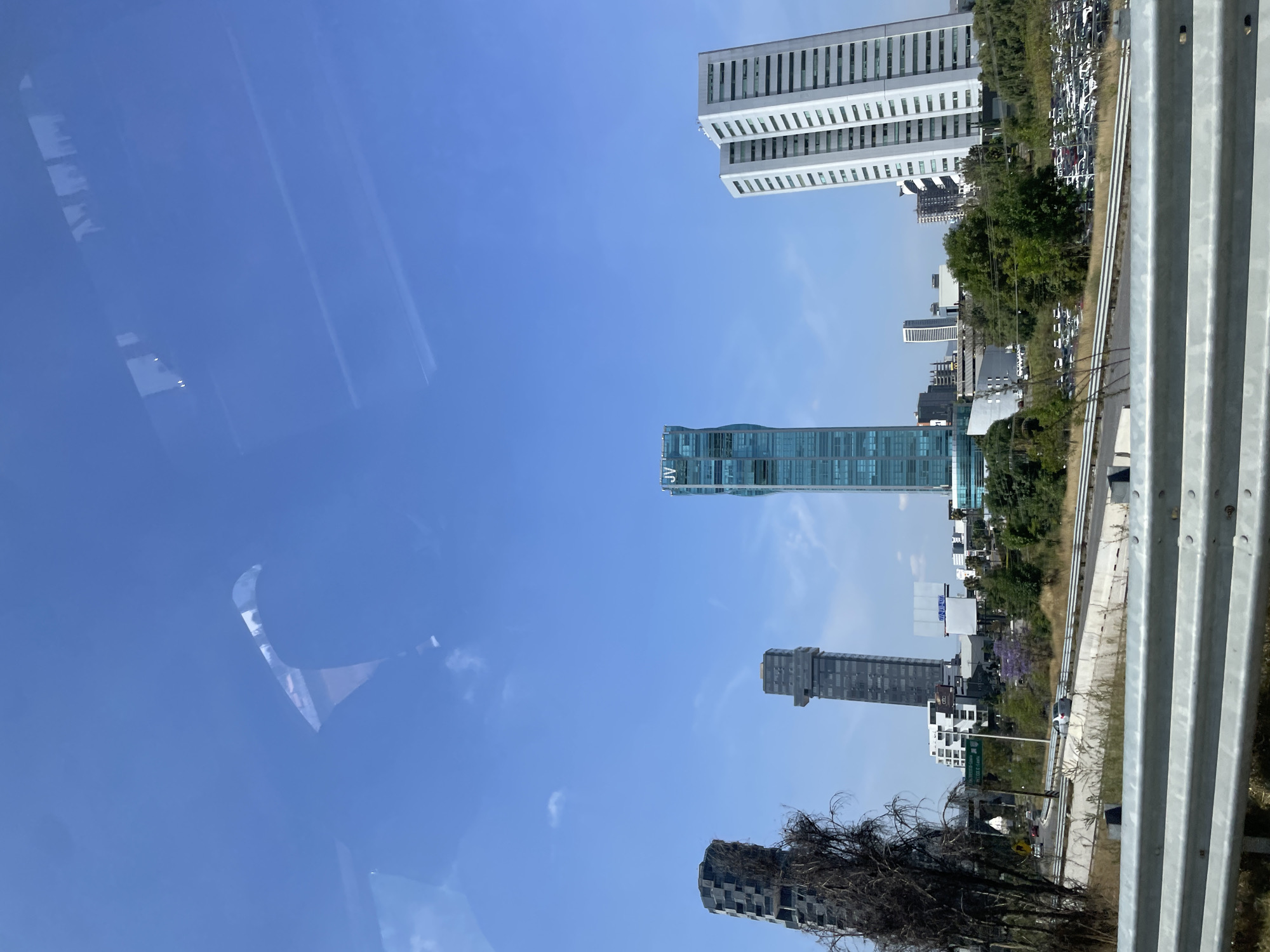 The image shows a view of the skyline in San Bernardino Tlaxcalancingo, Mexico, from a vehicle. The sky is blue with some clouds. Several tall buildings are visible, including a white skyscraper in the upper right, a blue-green glass building marked "JV" in the center, and other buildings in the distance. A metal guardrail is in the foreground on the right. The reflection of a person's face is visible in the upper left of the window. A row of parked cars is near the white skyscraper.
