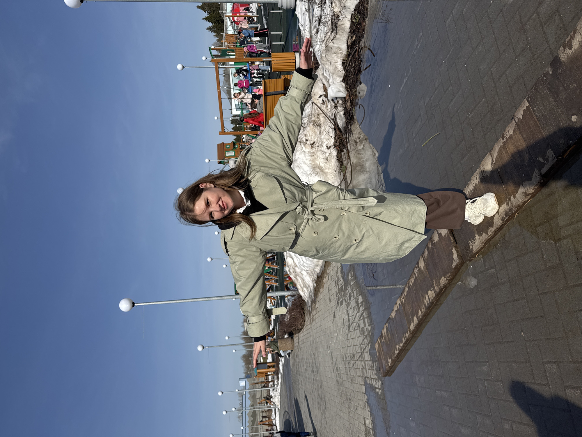 A young woman stands on a walkway in Perm, Russia, wearing a light green trench coat, brown trousers, and white sneakers. She has her arms outstretched. There is snow melting alongside the walkway, as well as playground equipment in the background. The sky is clear and blue with streetlamps in the distance. The walkway is made of gray paving stones.