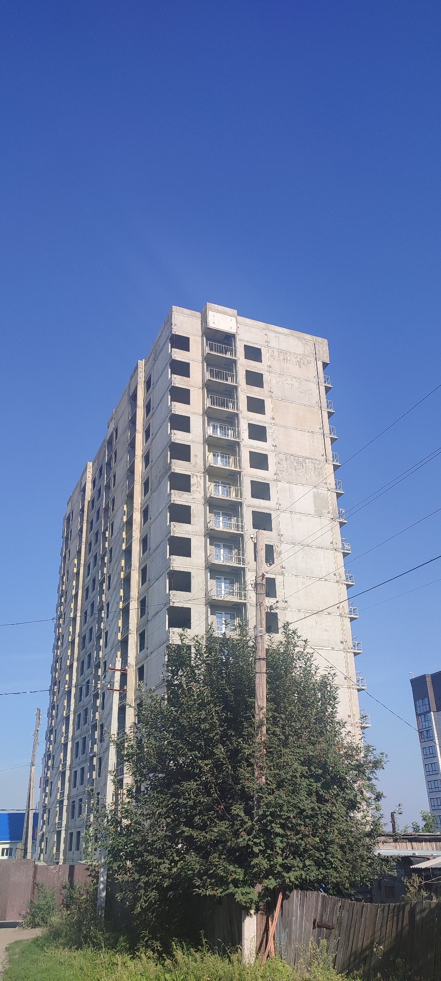 The image depicts an unfinished multi-story building under construction in Novoaltaysk, Russia. The building, made of concrete, has numerous square window openings and balconies with metal railings. Power lines are visible crisscrossing the sky. In the foreground, there is a large, dense green tree, a wooden utility pole, and a wooden fence. Adjacent to the unfinished building, a section of a completed building with a modern facade can be seen. The sky is clear and blue, indicating a daytime scene. The overall impression is one of urban development in progress, with contrasting elements of construction and existing infrastructure.