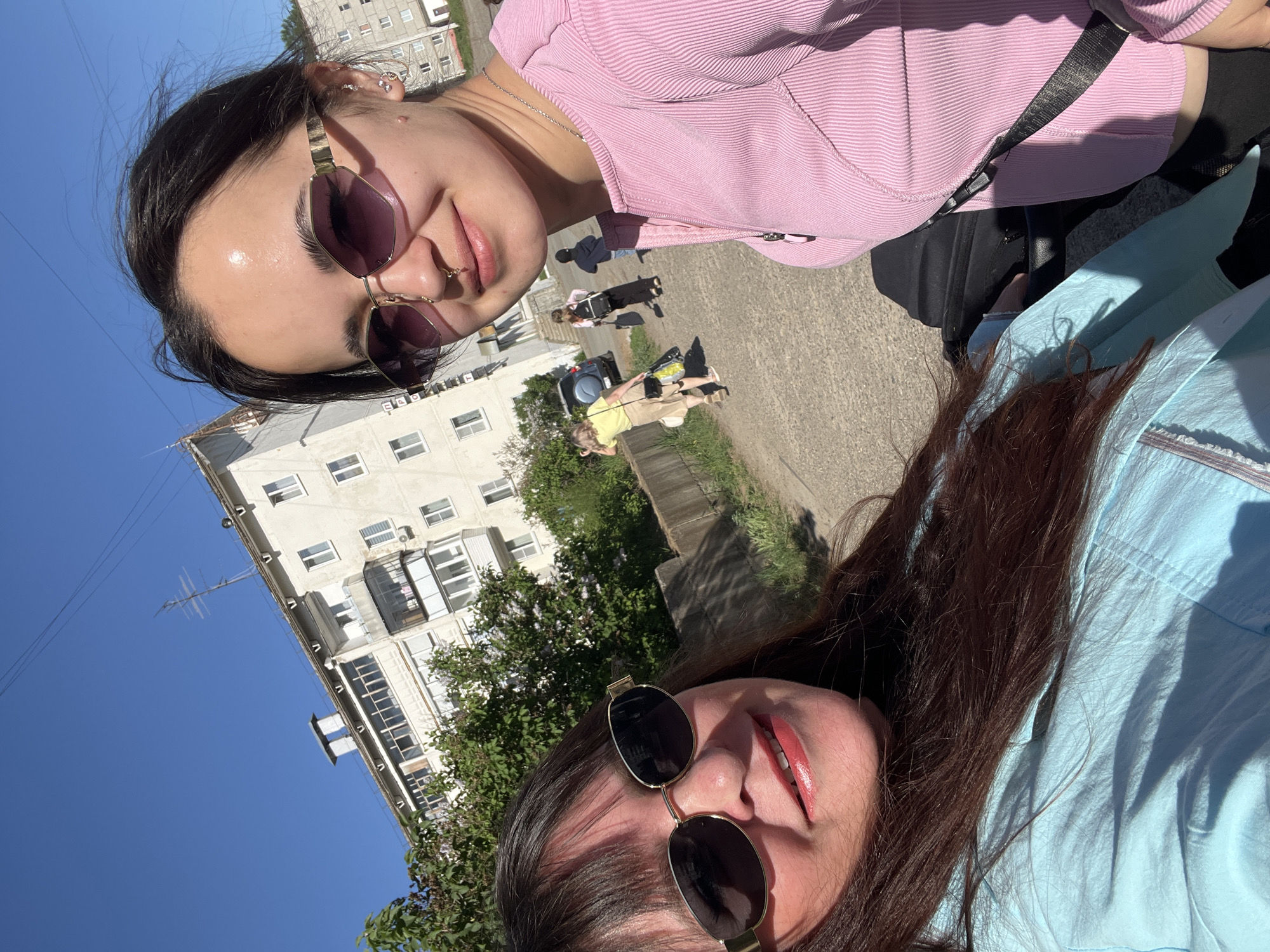 Two women are posing for a selfie on a street in Noviy Urengoy, Russia. One woman is wearing a pink top and sunglasses with a nose ring. The other woman wears a blue shirt and sunglasses. In the background, pedestrians walk on the sidewalk near a white apartment building.