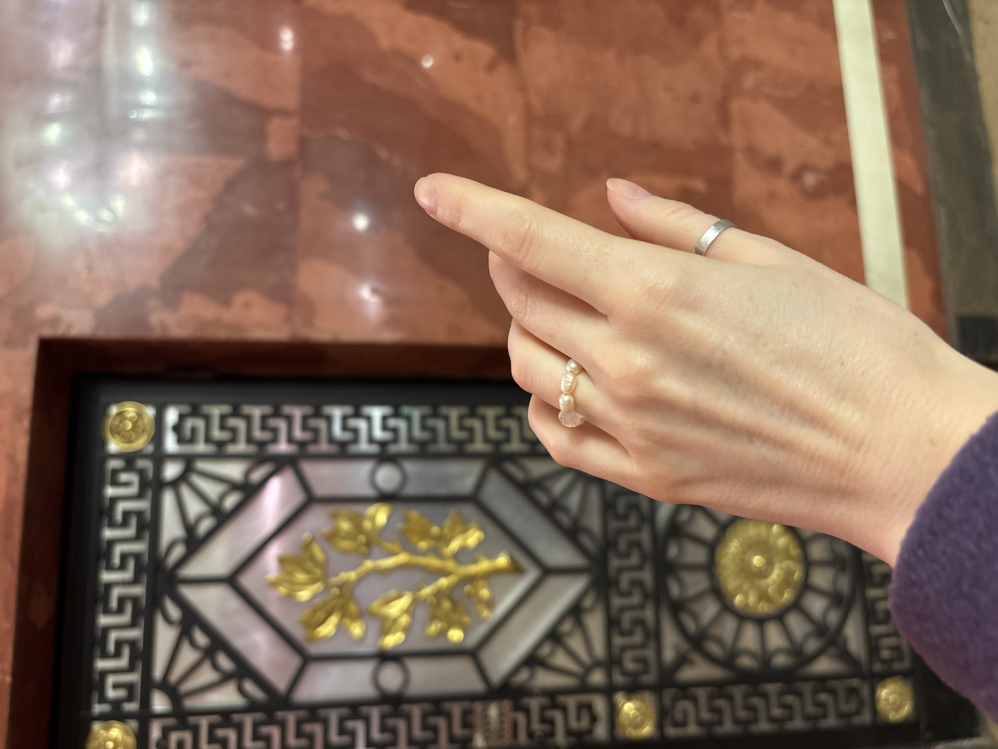 A close-up view in the Yusupov Palace on Moika Embankment, Saint Petersburg, Russia, shows a fair-skinned human right hand extending from the lower left. The hand's index finger points upwards and to the right. A ring featuring several irregularly shaped pearls is on the middle finger, and a plain silver-colored band is on the ring finger. The fingernails are natural and unpolished. The wrist is partially covered by a dark purple fabric. In the background to the left, a section of a dark-framed decorative panel or door is visible, featuring a central golden stylized tree or plant motif against an iridescent, possibly mother-of-pearl, background. This panel also includes black geometric patterns, golden circular embellishments, and a repeating Greek key border. To the right, a polished wall of reddish-brown marble or granite exhibits natural veining and bright reflections from overhead lighting.