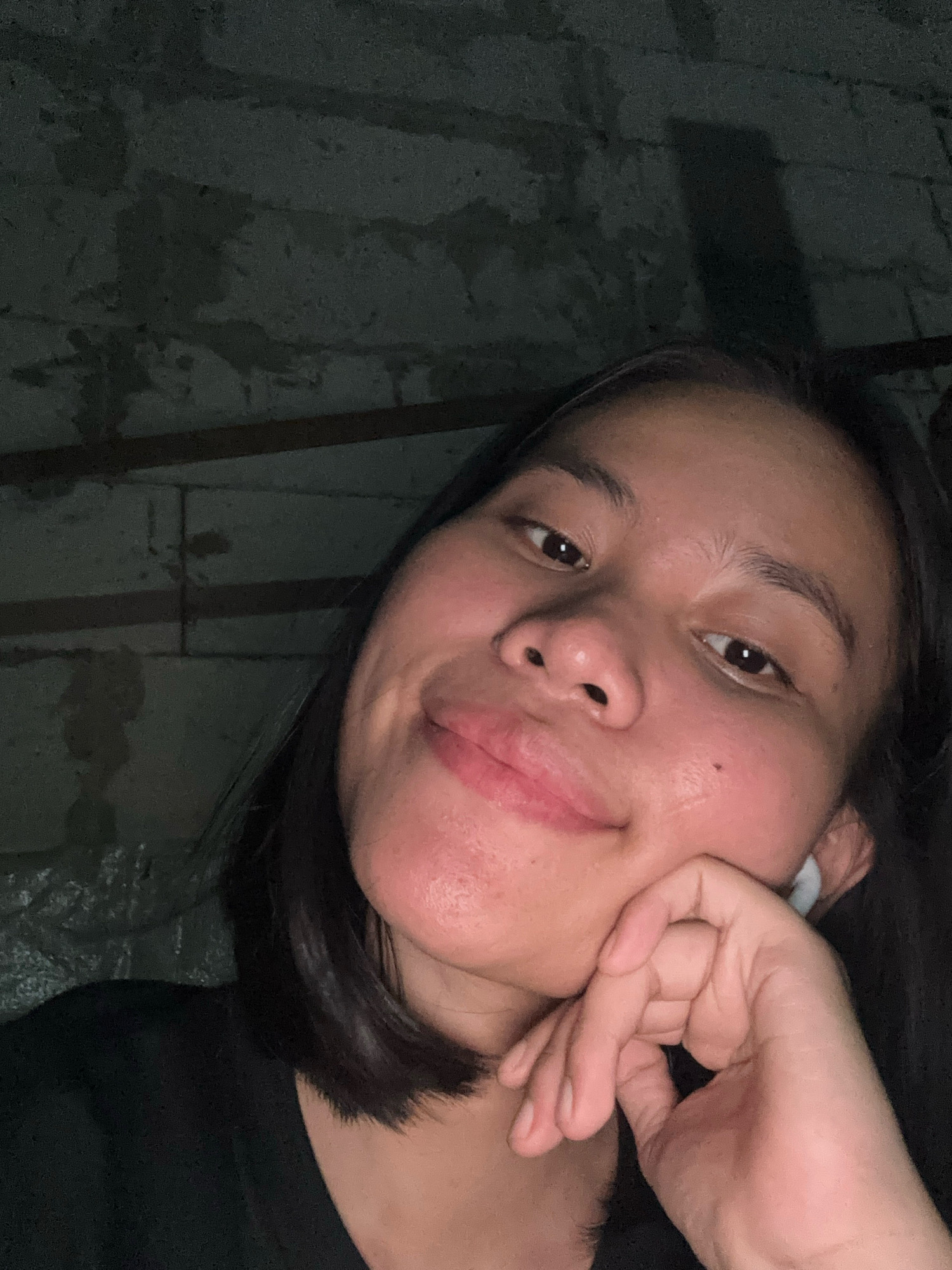 The image shows a close-up selfie of a young woman. She has dark hair, brown eyes, and is wearing a black t-shirt. Her left hand is resting on her cheek, and she is wearing a white earbud in her right ear. The background appears to be a rough, textured wall, possibly made of concrete blocks. The lighting is dim, casting shadows on the wall and highlighting the woman's face. The scene is indoors, with what appears to be wooden beams visible above the wall. There are no explicit indicators of location within East Jakarta, Indonesia, or any specific city.