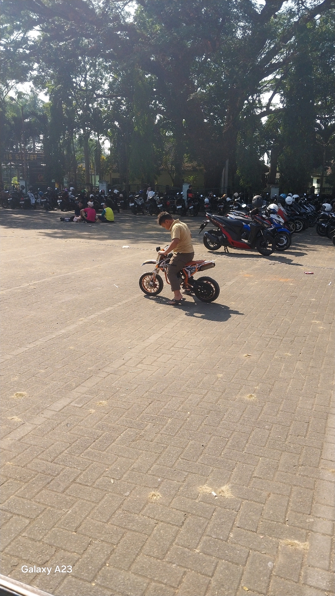 A man is sitting on a small dirt bike in an outdoor parking lot in Malang, Indonesia. The man, wearing a tan t-shirt and brown pants, is looking down. The dirt bike is orange and white. In the background, many motorcycles are parked, and several people are sitting on the ground near the motorcycles. Large trees with dense foliage are visible behind the parked motorcycles, and a building is partially visible. The ground is paved with interlocking bricks, and the sun is casting shadows. The image has a watermark in the lower left corner that reads "Galaxy A23".