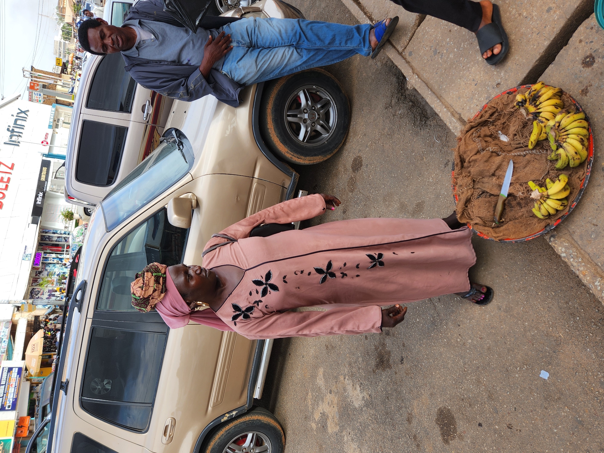The image captures a street scene in an unidentified city. A fair-skinned man, wearing a gray jacket, light blue jeans, and blue sandals, reclines on the hood of a beige SUV, his legs stretching over a front tire. Beside the SUV, a dark-skinned woman, dressed in a peach-colored dress with black floral accents, stands with a tray on the sidewalk. The tray contains bananas and a knife resting on a piece of brown burlap. A person wearing dark sandals and black pants stands next to the tray. Shops line the street, visible in the background, with signage indicating a commercial area.