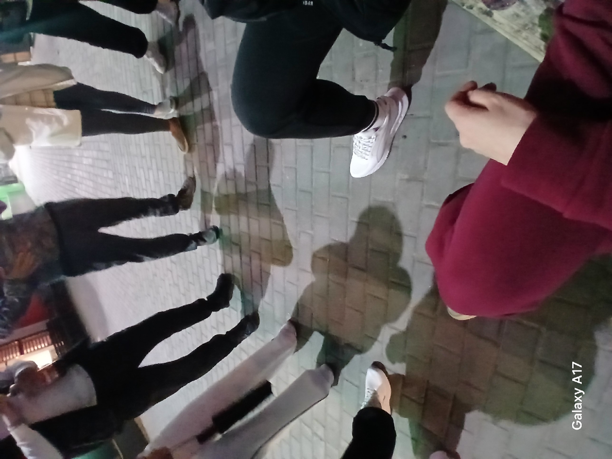 The image shows a group of people standing outside on a paved surface. The view is from above, looking down at their legs and feet. Several people are wearing dark pants and various types of shoes, including sneakers and boots. One person is wearing a maroon-colored garment. Shadows of the people are cast on the ground. The image includes a watermark "Galaxy A17". The image was taken in Yekaterinburg, Russia.