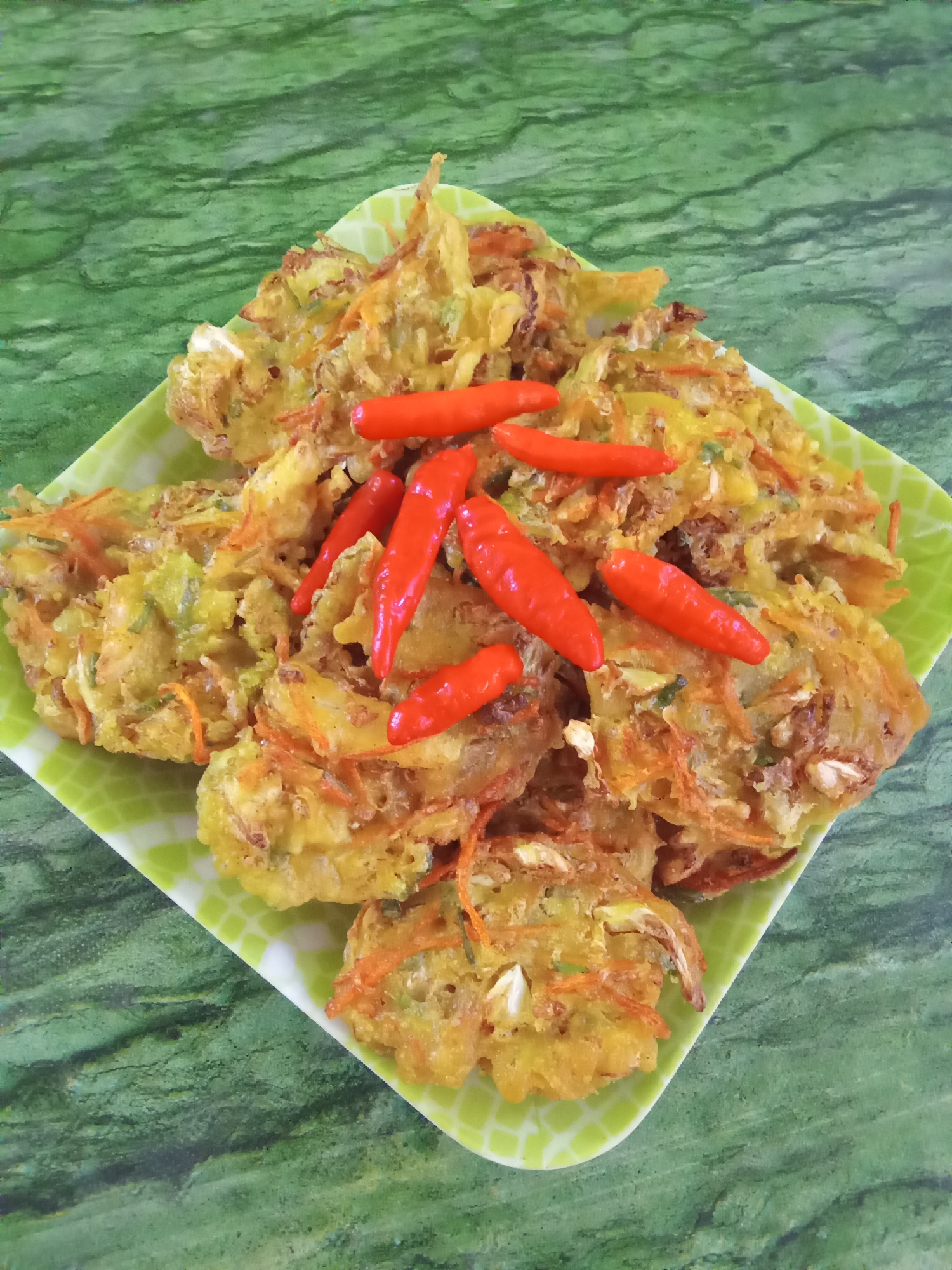 A plate of fried vegetable fritters, possibly bakwan sayur, is presented on a green, textured surface. The fritters are golden brown and appear to contain shredded vegetables such as carrots and cabbage, mixed with a batter. Several bright red chilies are placed on top of the fritters as a garnish. The plate itself is a light green color with a subtle geometric pattern around the rim. The background is a deep green with a marbled or wood-grain-like texture. The lighting suggests an indoor setting, possibly a kitchen or dining area in Subang, Indonesia. No people are visible in the image.