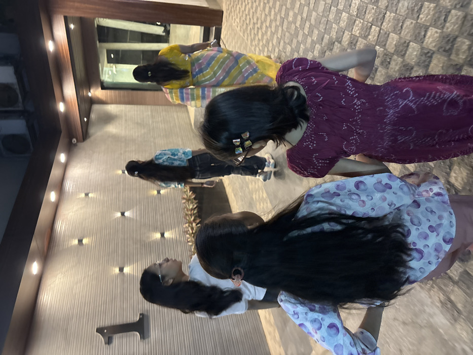 Four women stand in a row on a stone-paved surface, each facing away from the viewer. The woman closest to the camera wears a white shirt and light pink pants with a light purple, patterned jacket. The woman behind her is in a dark red dress. Following her is a woman in a blue top and black pants. Lastly, a woman in a yellow and multicolored dress is at the front. The background consists of a building with a light-colored, vertically striped wall, small light fixtures, and dark window panes.