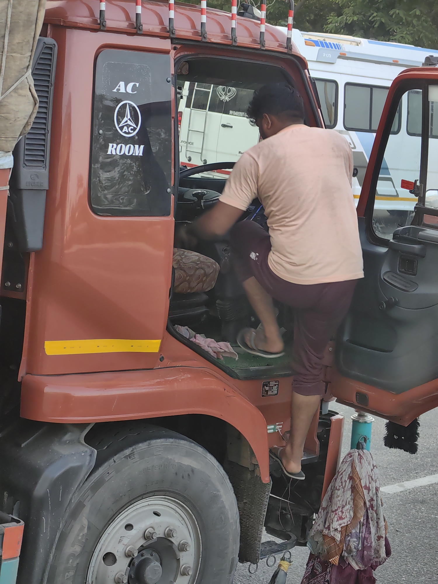 A man is stepping into the driver's seat of a red truck. The truck has "AC ROOM" written on the inside of the driver's side window. A white ambulance is parked behind the truck. The image was taken in Panipat, India.
