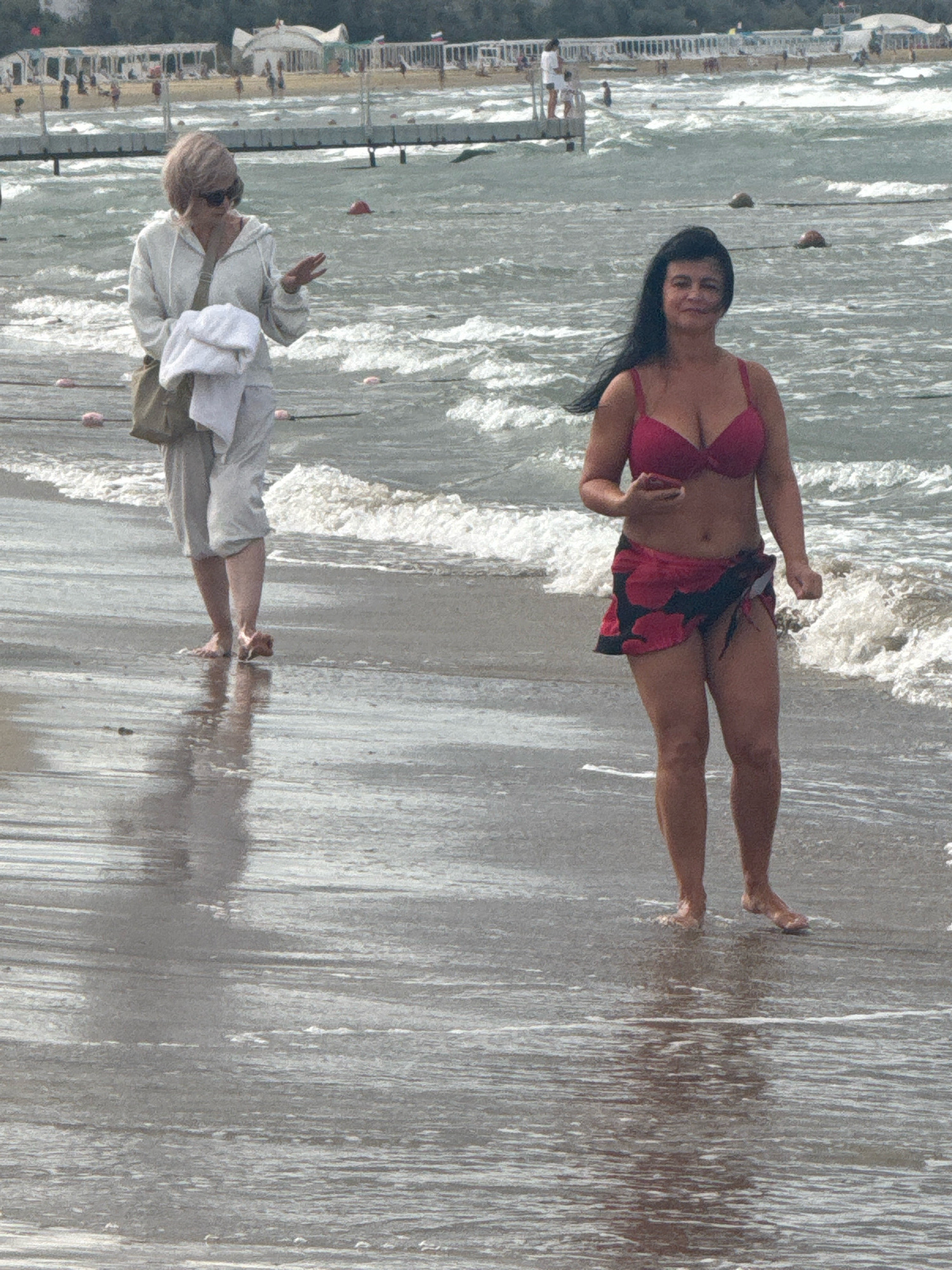 Two women are standing on a sandy beach. The woman on the left is wearing a light-colored hooded jacket, light-colored pants, sunglasses, and a light-colored purse across her shoulder. She is holding a white towel in her left arm and has her right hand raised. The woman on the right is wearing a red bikini top and a red and black floral sarong. She is holding a dark-colored object in her right hand and is looking at the camera. The background shows a body of water with waves, a pier with people, and some buildings in the distance. The scene appears to be a public beach in Anapa, Russia.