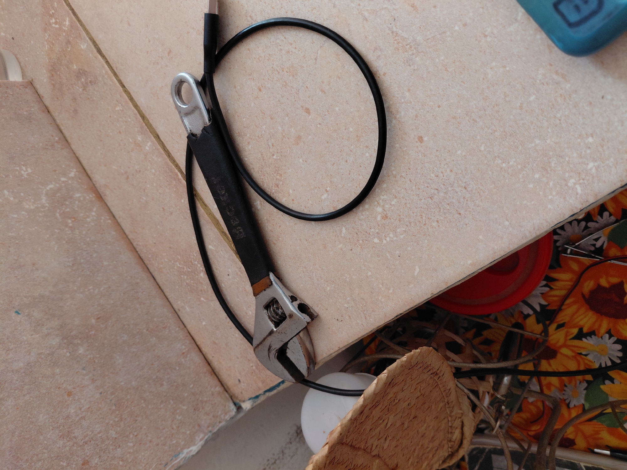 An adjustable wrench and a black cable rest on a light brown tiled surface, with the scene set in Santa Ana, Venezuela. The metallic wrench features a black handle, where the white text "SEC KEY" is clearly visible, alongside other fainter, illegible letters. The black cable, which appears to be a charging cable with a silver-colored USB connector at one end, is coiled into a loop and wrapped around the wrench.
The tiled surface is speckled with lighter and darker brown tones and has visible grout lines. In the upper right background, a partial view of a light blue plastic object can be seen. The lower right portion of the image reveals a vibrant floral fabric patterned with yellow sunflowers and white daisies, along with a red object, possibly a bowl or lid, and a tangle of wires or cables. In the lower-left foreground, a light brown woven object, resembling part of a basket or mat, is partially visible. No specific activity is taking place; the items are stationary. The overall setting appears to be a functional or utility space, possibly a workshop or a cluttered counter.
