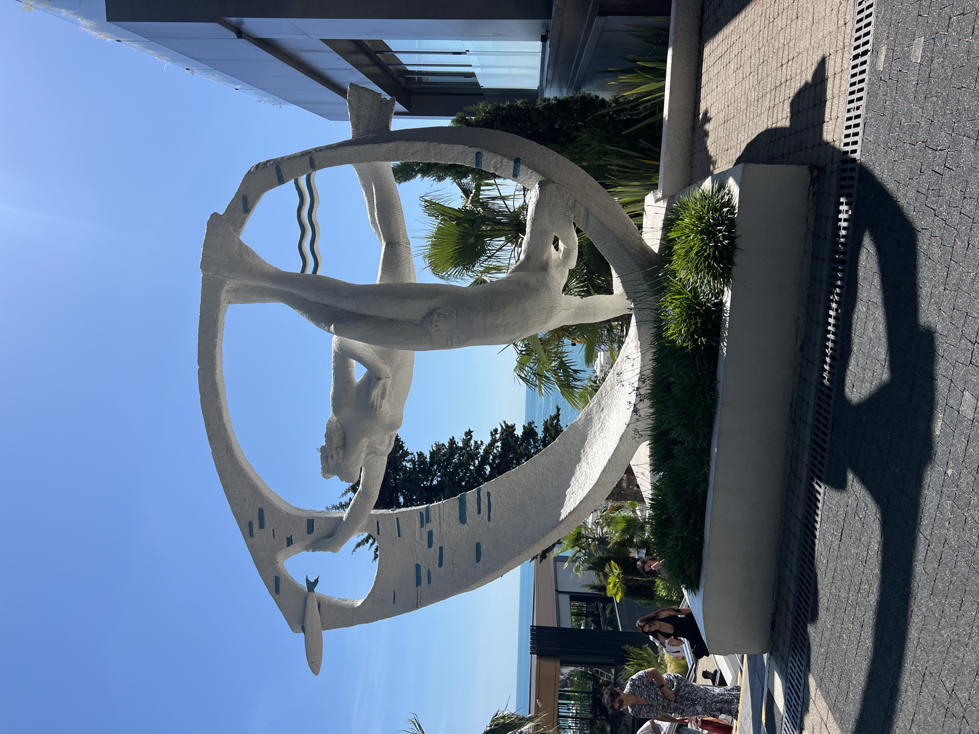 This image captures a large, abstract white sculpture depicting a figure swimming or diving within a circular frame, set against a bright blue sky. The sculpture is positioned outdoors, likely in a public area. Several people are visible on a set of stairs in the background, some walking up and others descending. To the right of the sculpture is a modern building with glass windows. Palm trees and other greenery are scattered around the scene, and the ocean is visible in the distance, suggesting a coastal location. The shadow of the sculpture and possibly a person are cast on the paved ground in the foreground.
This sculpture is located at the "The Pearl of the Caucasus" sanatorium in the Khostinsky District of Sochi, Russia. The sculpture is titled "Diver" and was created by sculptor Lev Yakovlevich Karavatsky.