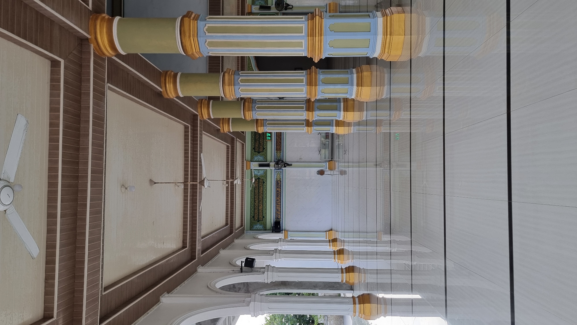 The image depicts the interior of a mosque in Meunasahlhok, Indonesia. The perspective is low, looking down the length of a hallway lined with ornate columns. The floor is highly polished, reflecting the white and gold columns and the ceiling above. The ceiling is coffered with alternating panels of wood and a cream-colored material, with several white ceiling fans installed.
Along the left side of the hallway, a series of white arched colonnades are visible, leading to an outdoor area with some greenery. On the right side, the hallway is defined by large, decorative columns painted in alternating sections of gold, light blue, and a muted green. These columns are the most striking architectural feature.
In the background, the wall of the prayer hall is visible. It features Arabic calligraphy inscribed on a green backdrop, illuminated by what appears to be fluorescent lighting. A digital clock displaying "17 37" is mounted on the wall. Two black oscillating fans are mounted on the wall below the calligraphy. The overall impression is one of a clean, well-maintained, and architecturally interesting religious space. No people are visible in the image.