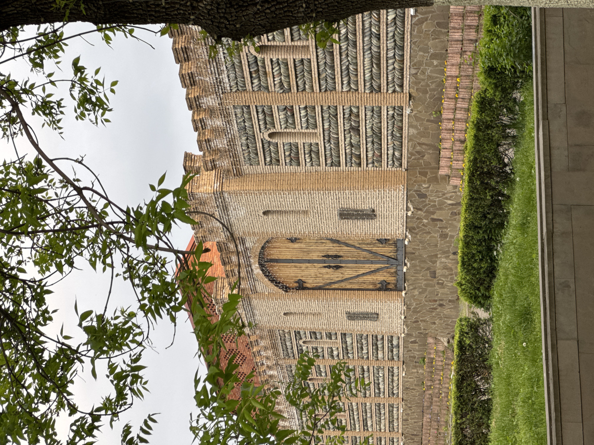 The image captures a section of a building located in Akhieli, Georgia. The structure is composed of brick and stone, with notable decorative brickwork. A wooden door with metal hardware is visible. A portion of a tree with green leaves is in the foreground, partially obscuring the building. Adjacent to the building is a landscaped area featuring greenery and a stone pathway. The sky is overcast.