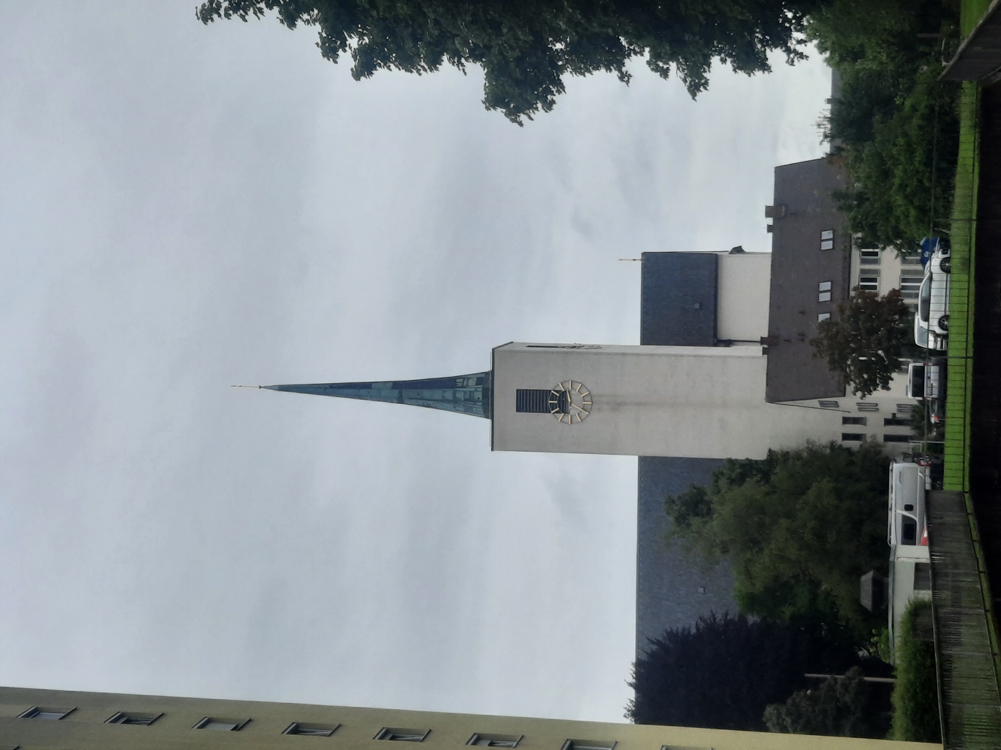 The image shows a tall, white church tower with a green spire and a clock face. In front of the church, there is a darker building with a dark grey roof. To the left of the church is a tall, light-colored building with many windows. In the foreground, there is a wooden railing with a canal or body of water beyond it, and grass and trees lining the bank. There are several vehicles visible in front of the buildings, including a white van and a white car. The sky is overcast.