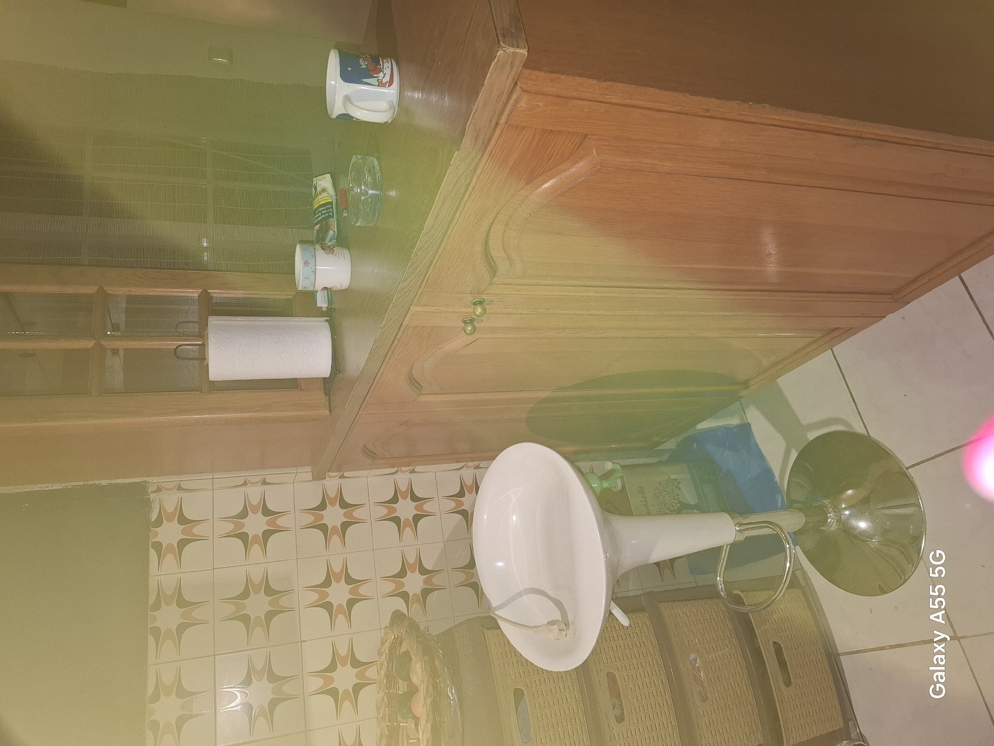 This image shows a corner of what appears to be a kitchen or bar area. A white bar stool with a chrome base is positioned in the foreground. Behind the stool, there are several woven storage baskets stacked. To the right, a wooden cabinet with two drawers is visible, topped with a wooden counter. On the counter, there is a roll of paper towels, two mugs, and an ashtray with a cigarette. The wall behind the counter is tiled with a retro-style pattern in beige, brown, and light orange. The lighting in the image has a strong green and yellow tint. There are no people or specific landmarks indicating the location within the Municipal Unit of Dafni, Greece. The "Galaxy A55 5G" watermark is visible in the bottom left corner.