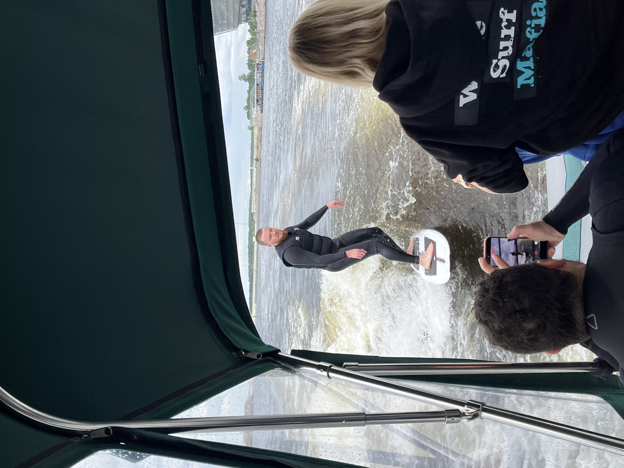 In this image, a group of people are on a boat on a body of water, likely a river or canal, in Saint Petersburg, Russia. The sky appears overcast, suggesting a cloudy day. The primary activity is wakeboarding, with a man in a black wetsuit actively riding a wakeboard behind the boat. His body is angled as he balances on the board, creating a spray of water.
In the foreground, a person with blonde hair, wearing a black hoodie with white and blue text, is looking out at the wakeboarder. The visible text on the hoodie reads "Surf Mafia". Another person, partially visible from behind, is holding up a smartphone, presumably to take a photo or video of the wakeboarding action. This person is also wearing a black wetsuit. The boat itself appears to be a pleasure craft with a dark green canopy providing shade, and metal railings in the foreground. The background shows a cityscape with buildings and infrastructure along the waterfront, consistent with an urban environment like Saint Petersburg. The overall impression is of an active outdoor recreational pursuit on a day with less than ideal weather.