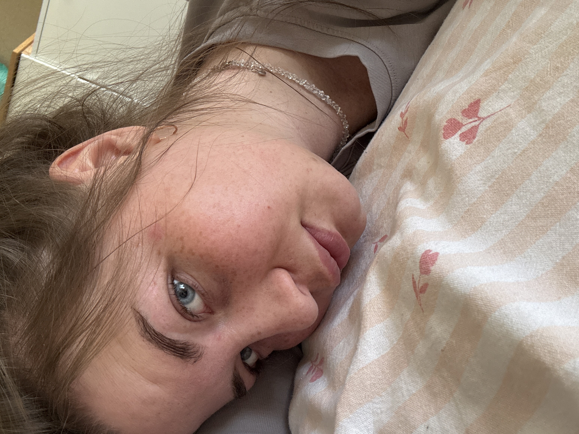 A close-up, slightly angled shot depicts a woman lying on a bed, positioned on her side. She has fair skin, light brown hair, and striking blue eyes. Freckles are visible on her nose and cheeks. She is wearing a light grey top and a delicate silver necklace around her neck. The bed is covered with a peach and white striped pillowcase featuring a floral pattern. The location is not discernible and cannot be determined to be in Chaikovsky, Russia, based on the visual information provided.