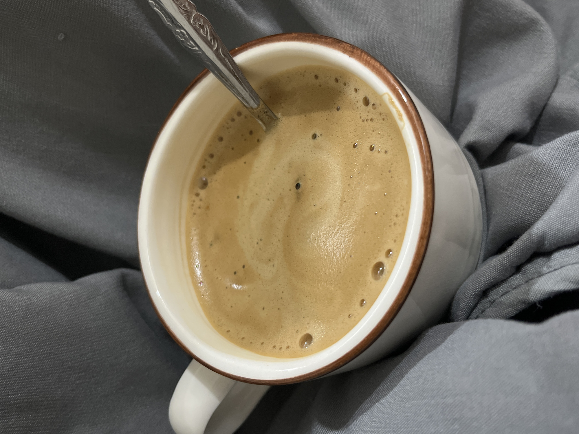 A close-up, overhead shot shows a white mug of coffee with a brown rim, filled with frothy coffee and a spoon resting in it. The mug is placed on a grey, textured surface, possibly a fabric. The background is a blur of grey fabric, suggesting a soft, draped material. The lighting is dim, highlighting the rich color of the coffee. No people are visible in the image. The location is not identifiable from the image itself.