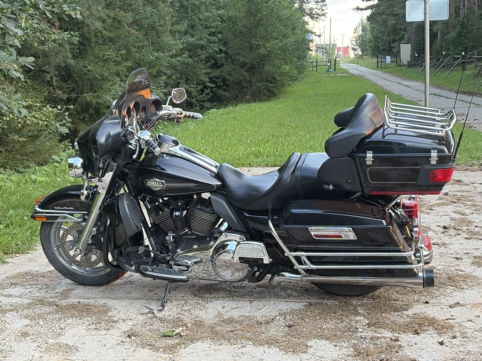 A black Harley-Davidson Ultra Classic motorcycle is parked on a dirt and gravel path. The motorcycle is facing left and is viewed from the side. The motorcycle has a large windshield, a black leather seat, and saddlebags. A chrome luggage rack is mounted on the back of the motorcycle. The motorcycle is surrounded by trees and grass. The sky is overcast and the time of day appears to be late afternoon. There are no people in the image. The text "HARLEY-DAVIDSON" is visible on the fuel tank, and "Ultra Classic" is visible on the front fender.