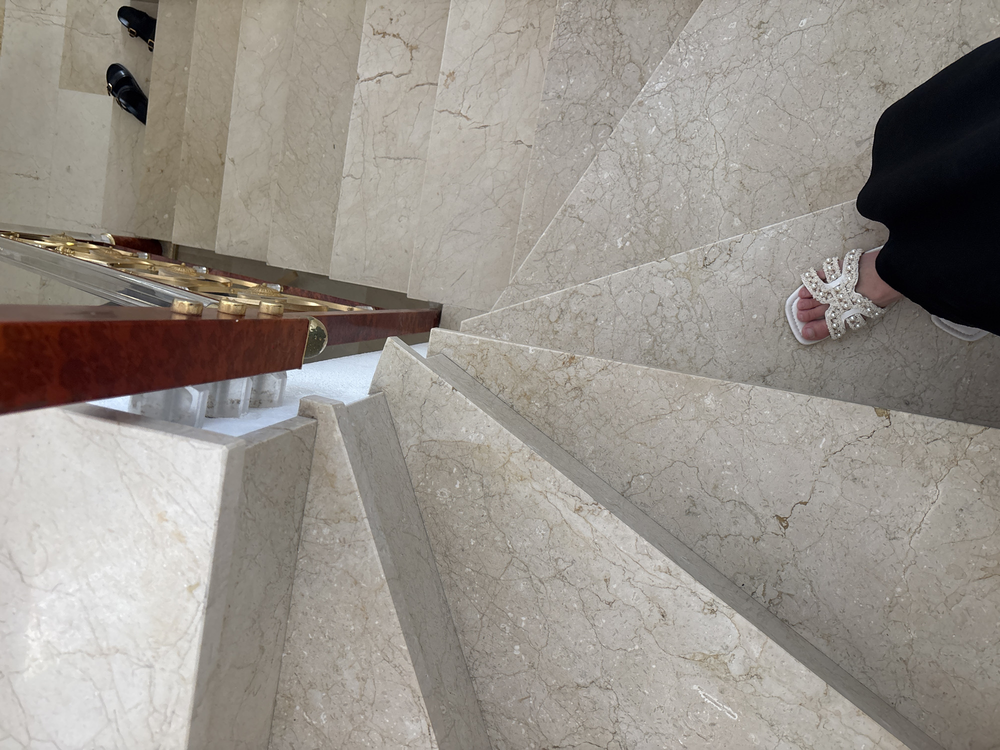 The image shows a marble staircase with a wooden and brass handrail. On the stairs, there is a person's foot wearing a white sandal adorned with pearls. Further up the stairs, a pair of black sandals is visible. The location is stated to be Orihuela, Spain.
