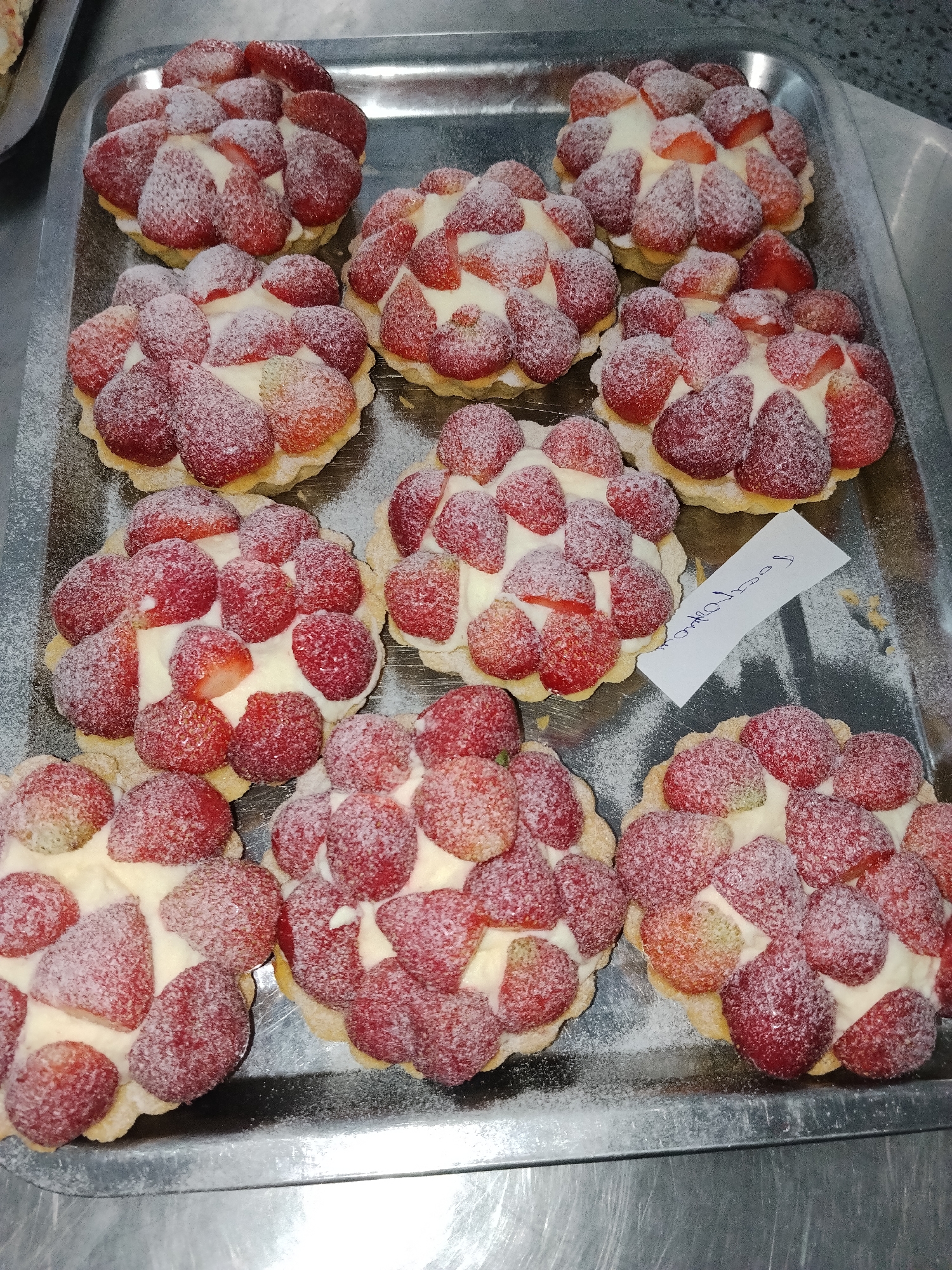A tray of ten strawberry tarts dusted with powdered sugar is presented on a silver metallic tray. The tarts are arranged in three rows: the top row has three tarts, the middle row has four, and the bottom row has three. Each tart features a pastry shell filled with a white cream and topped with numerous halved or quartered strawberries. A small white piece of paper with handwritten text in an unfamiliar script is placed on one of the middle tarts. The background suggests an indoor setting, possibly a bakery or food preparation area, with hints of other food items and a countertop visible. There are no people in the frame. The lighting is artificial, casting some shadows. The scene is clean and organized. The location is identified as Bishkek City, Kyrgyzstan.