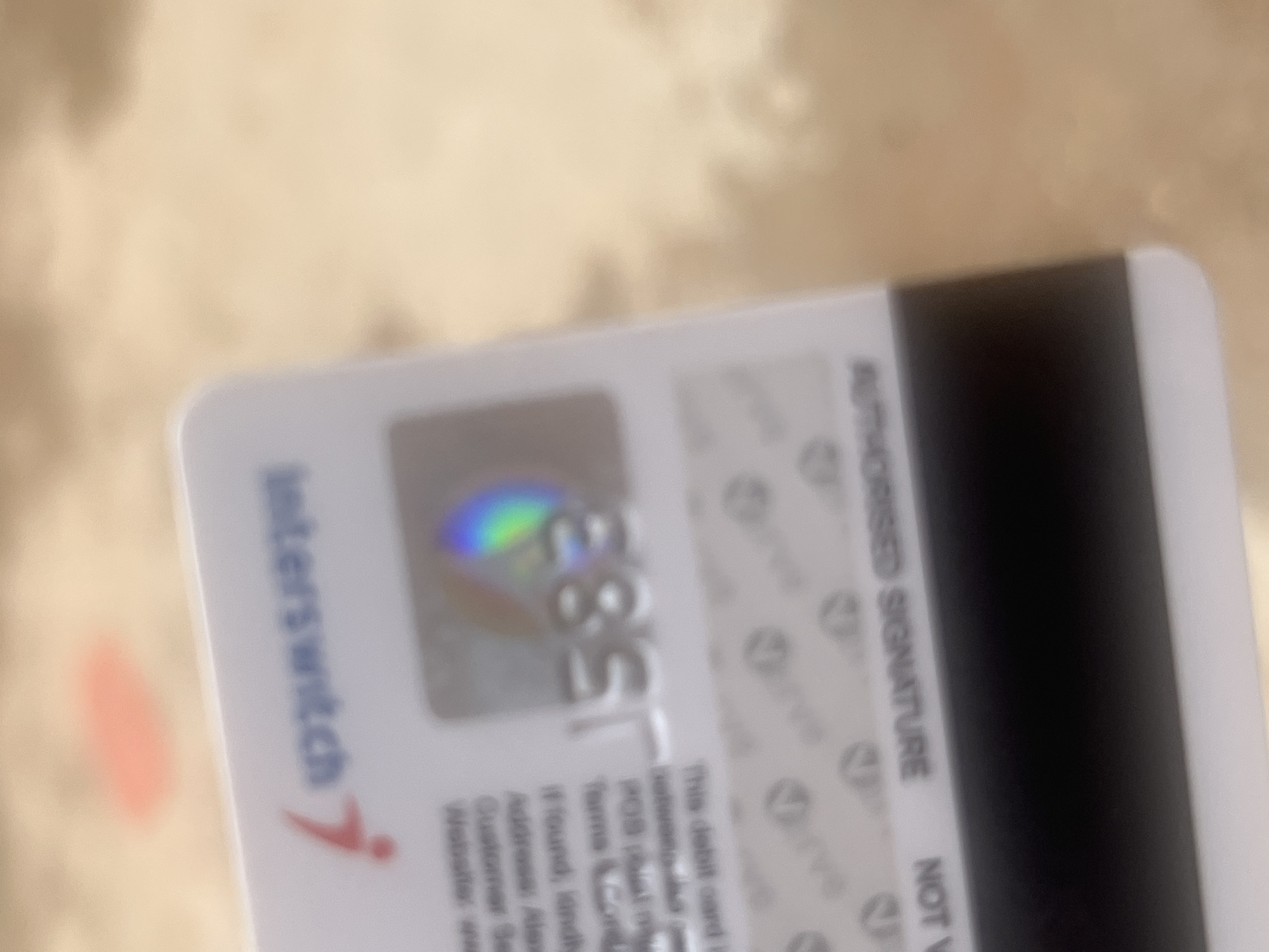 This is a close-up, blurry photograph of the back of a debit card. The card is white with black magnetic strip on the right side. Text and logos are visible on the white surface. The text "Interswitch" is printed vertically on the left side of the card, along with a red checkmark logo. A holographic security feature with rainbow colors is also visible. Below this, there are lines of text, partially obscured and difficult to read due to the blurriness, but some words like "AUTHORISED SIGNATURE", "NOT V", "This debit card", "Terms & Cond", "If found landly", "Customer Se" are discernible. The background is a blurred, textured surface that appears to be a light brown or beige color, possibly a table or a wall. The lighting suggests it might be indoors or in a shaded area, as there are no strong shadows. The photograph is taken at an angle, with the card diagonally positioned within the frame. There are no people visible in the image. The overall impression is that of a quick, informal photo of a debit card, likely for identification or documentation purposes. The provided location context of Jalingo, Nigeria, does not offer specific visual cues within the image itself but places the origin of the card within that region. No time of day or weather information can be deduced from the image. The emotions conveyed are neutral, as the focus is solely on the object.