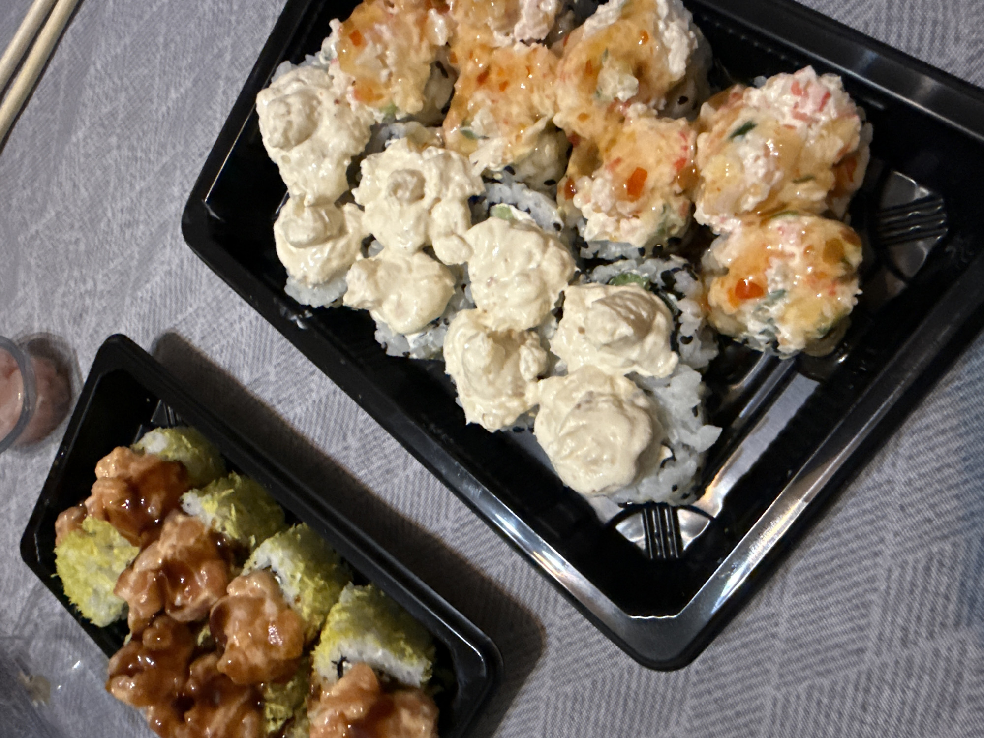 Two black plastic takeout containers are arranged on a textured gray surface. The larger container on the right holds sushi rolls topped with a light cream-colored topping and a mix of cream and orange toppings, each with a drizzle of sauce. The smaller container on the left contains sushi rolls, some coated in green and others with a brown topping with a drizzle of sauce. Chopsticks rest to the left of the containers. A small container possibly containing sauce, sits next to the smaller container. The scene suggests a meal of sushi. There is no evidence to identify the city of Varna, Bulgaria.