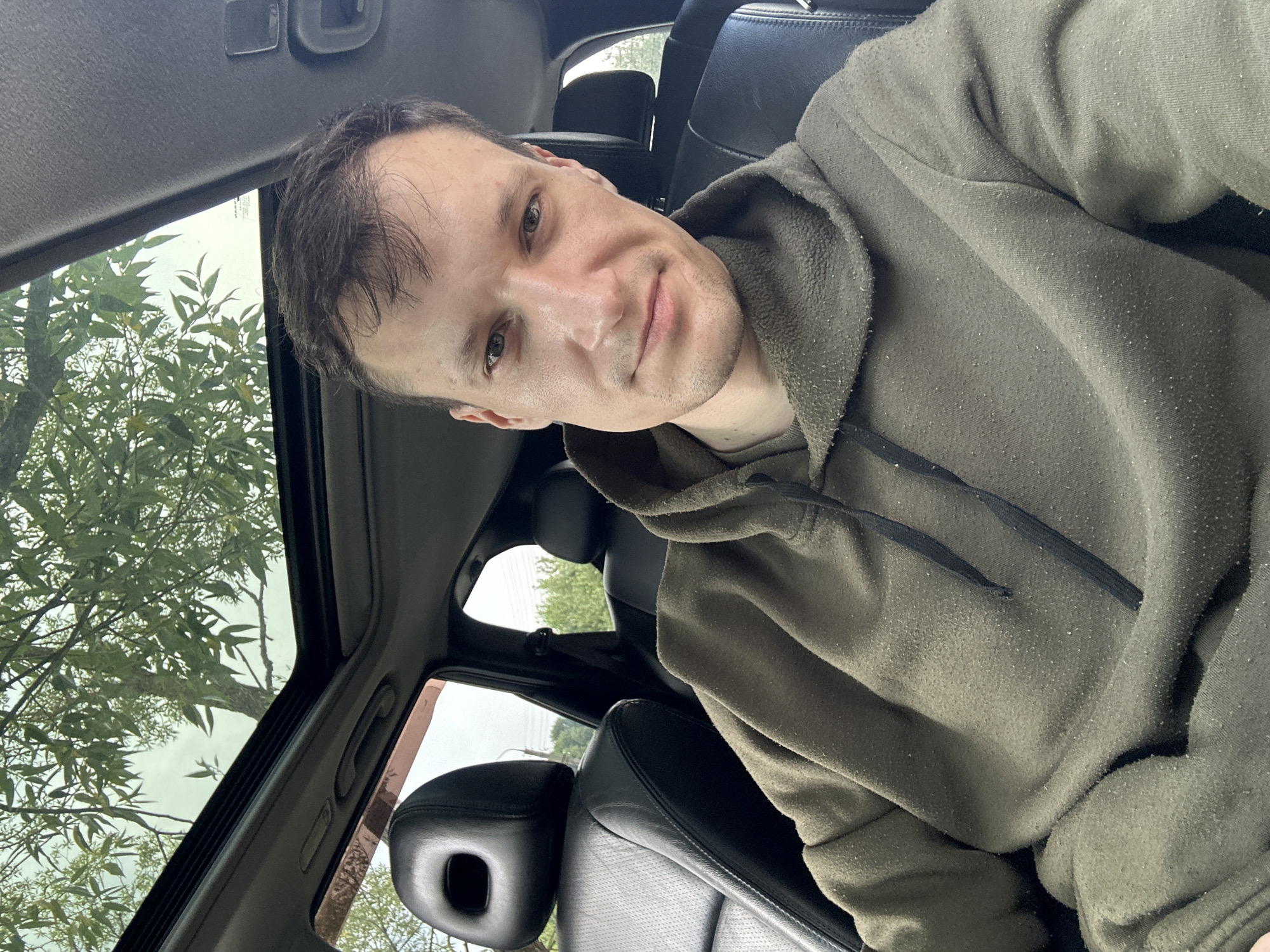 A man is sitting inside a car with the sunroof open, looking at the camera. He is wearing a green hooded sweatshirt. The car's interior is visible, including black leather seats and a portion of the dashboard. Outside the car, green trees and foliage can be seen through the open sunroof and windows. The image appears to have been taken outdoors during the daytime, possibly in a rural or suburban setting outside the city of Makiv, Ukraine.