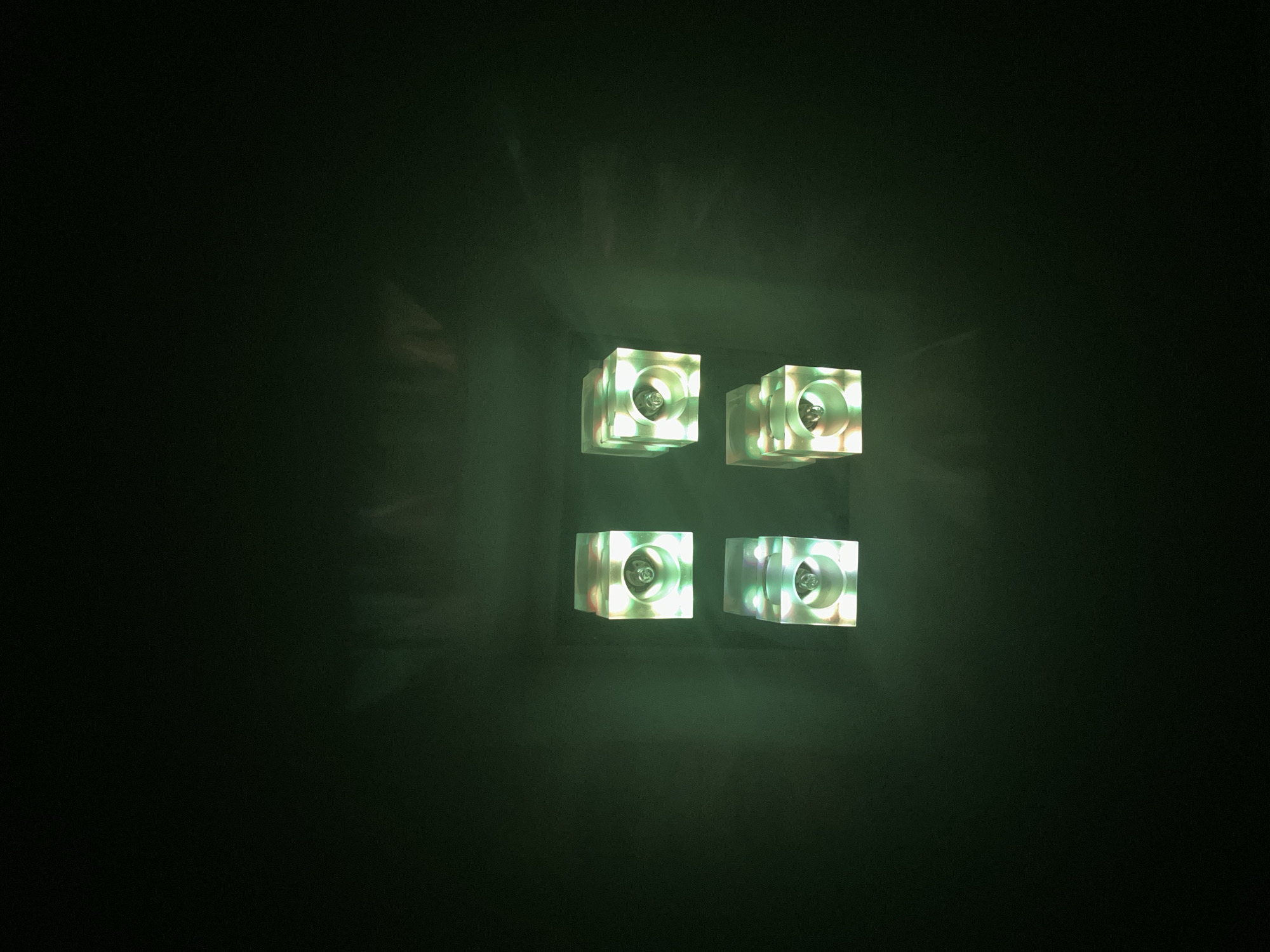The image shows four cube-shaped lights arranged in a 2x2 grid against a dark, slightly textured background. Each cube appears to be made of a translucent material and emits a soft, multicolored light, primarily in shades of green, white, and pink, with a central illuminated element. The lights are positioned in the upper-middle section of the frame, with the background fading into darkness at the top and bottom. There are no people, animals, or other objects visible in the scene. The lighting suggests a dimly lit environment. This image does not provide any contextual information about its location in the city of Warsaw, Poland.